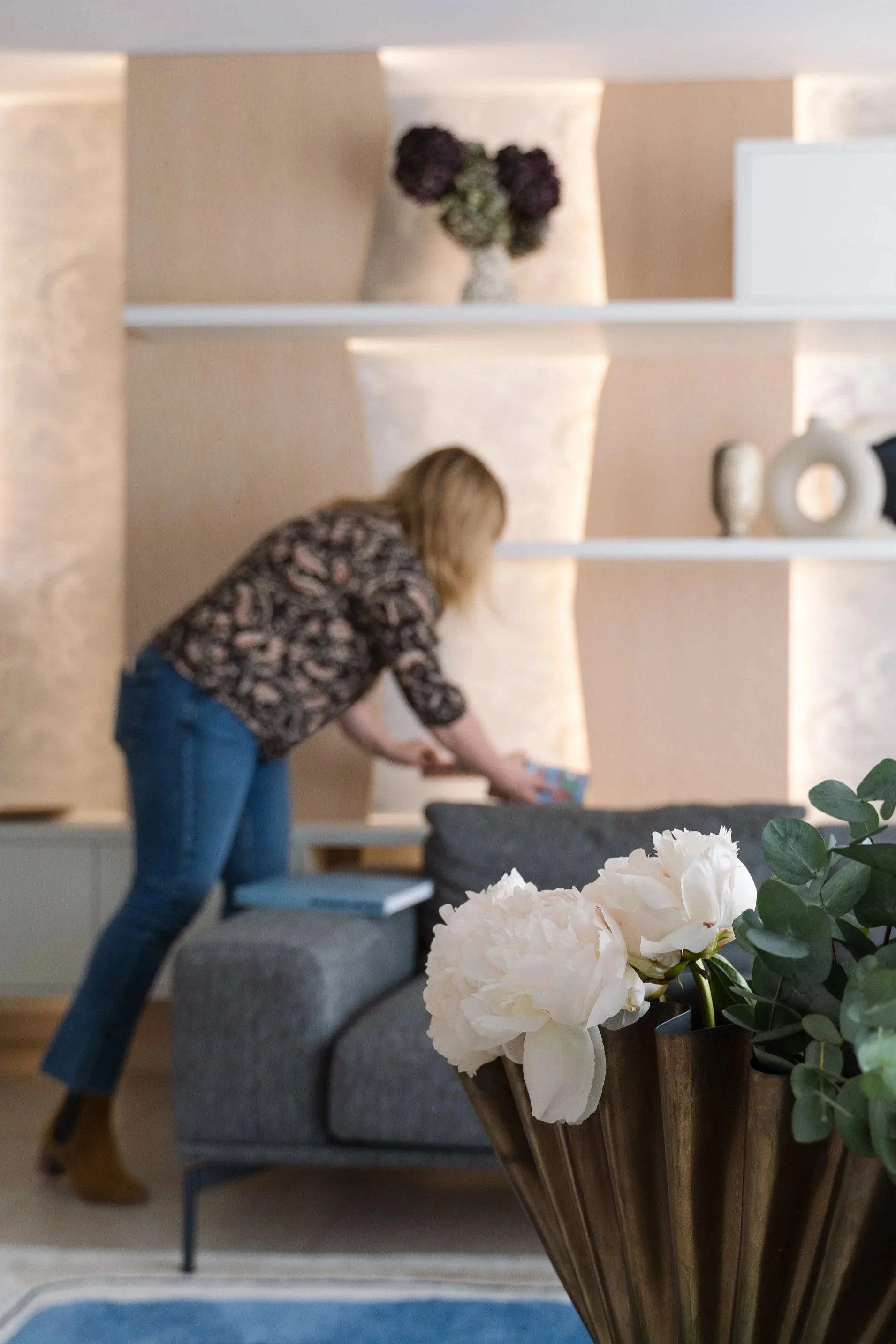 mélanie Trinkwell, designer d'intérieur, arrangeant un livre sur une table. Dans le premier plan, il y a un grand vase avec des fleurs blanches et des feuilles vertes. Le salon a un mur en bois clair avec des étagères décoratives.