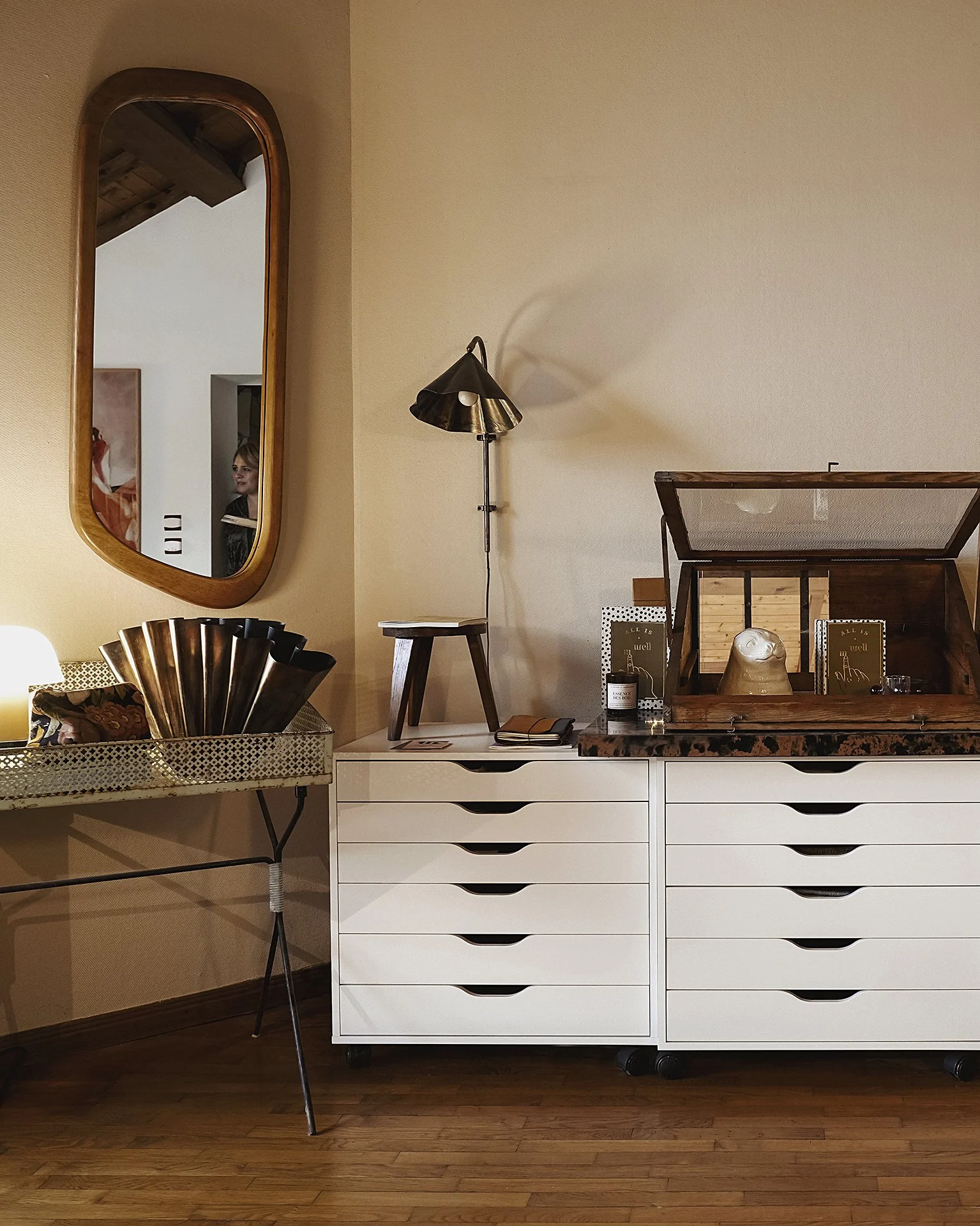 Intérieur d'une pièce avec un grand miroir ovale en bois accroché au mur, une lampe de lecture noire à côté, une commode blanche à dix tiroirs, un petit bureau avec une chaise en métal, certains objets décoratifs et livres, un mur beige et un sol en bois.