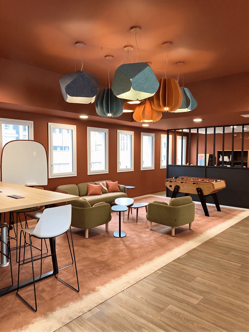 espace coworking CPAM avec canapé, fauteuils, tables basses, tapis rose, lampes suspendues colorées, vue sur plusieurs fenêtres et une aire de jeux avec baby-foot.