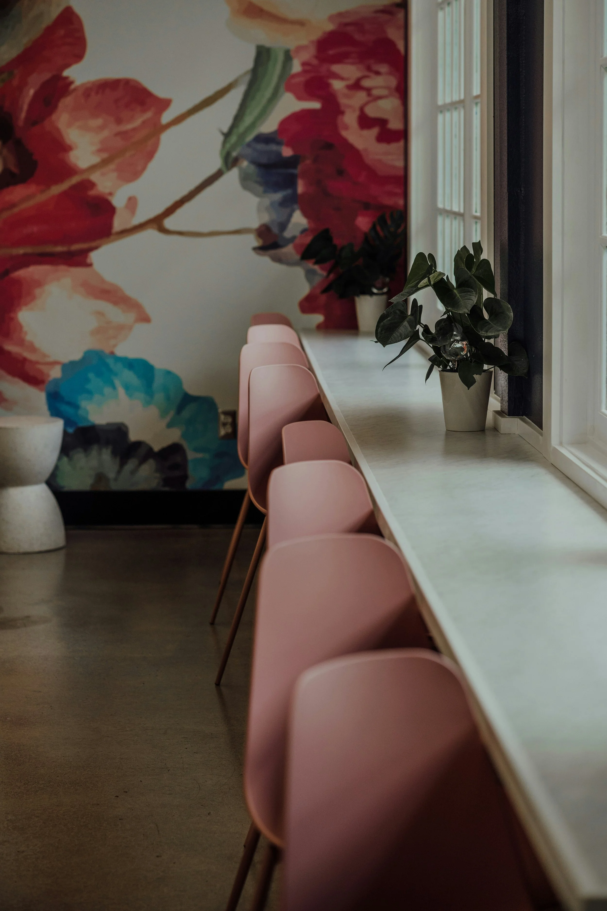 Une vue d'une table de cafe avec des chaises roses, une plante en pot et un mur décoré d'une peinture florale
