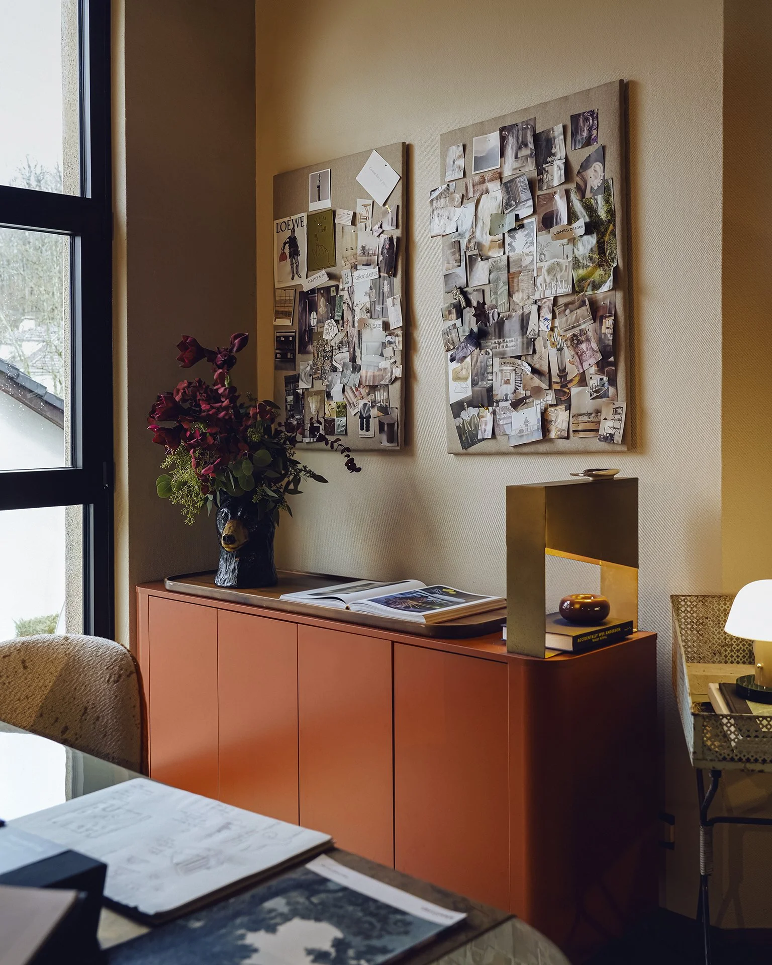 Une pièce intérieure avec un meuble orange foncé, un bouquet de fleurs violettes dans une tête de chien sculptée en bois, et deux panneaux de liège recouverts de photos et de papiers. Un livre ouvert repose sur la surface, et une fenêtre laisse entrer de la lumière naturelle.