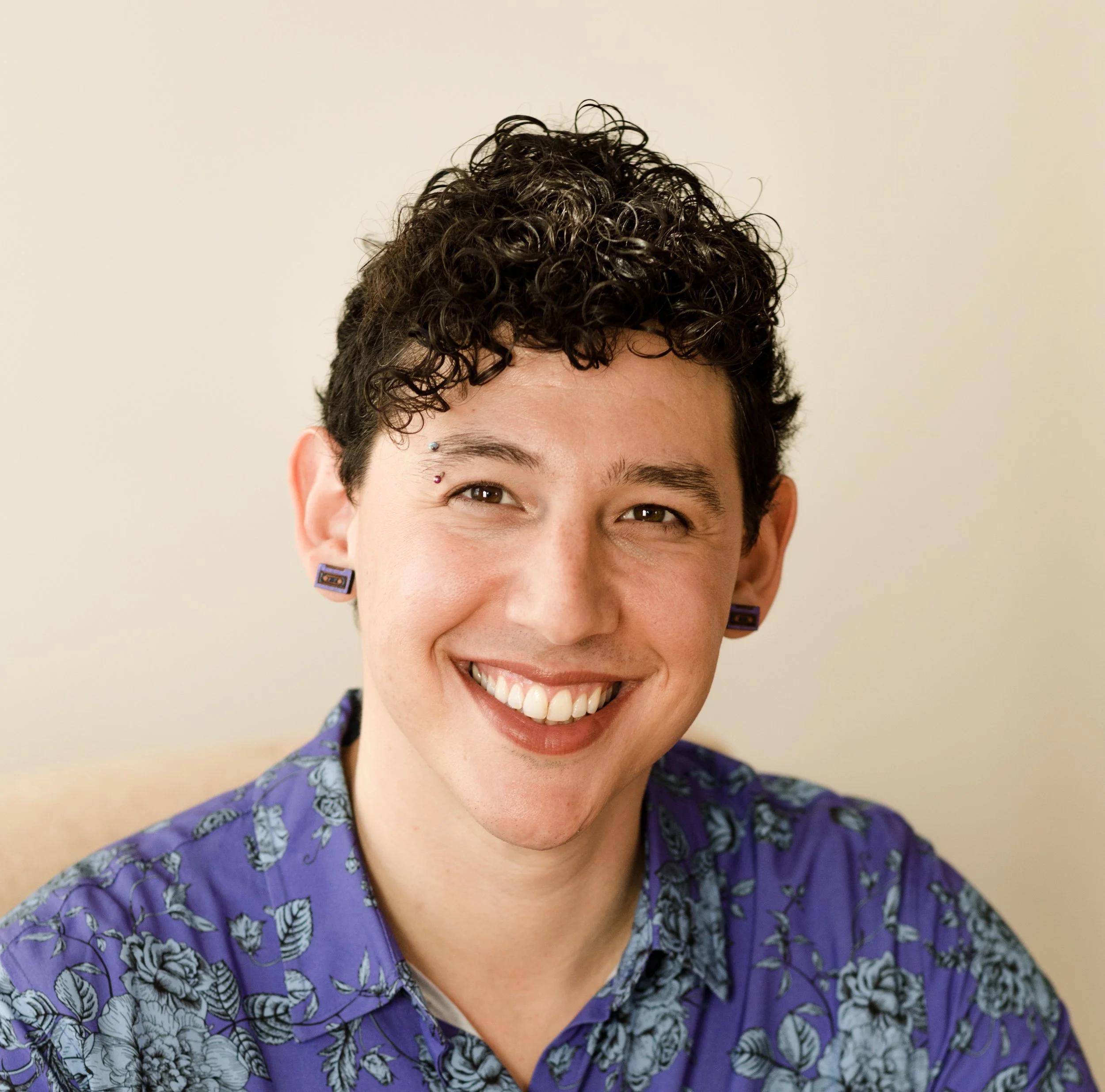 Close-up of a smiling person with curly dark hair, piercings, purple earrings, and a purple floral shirt.