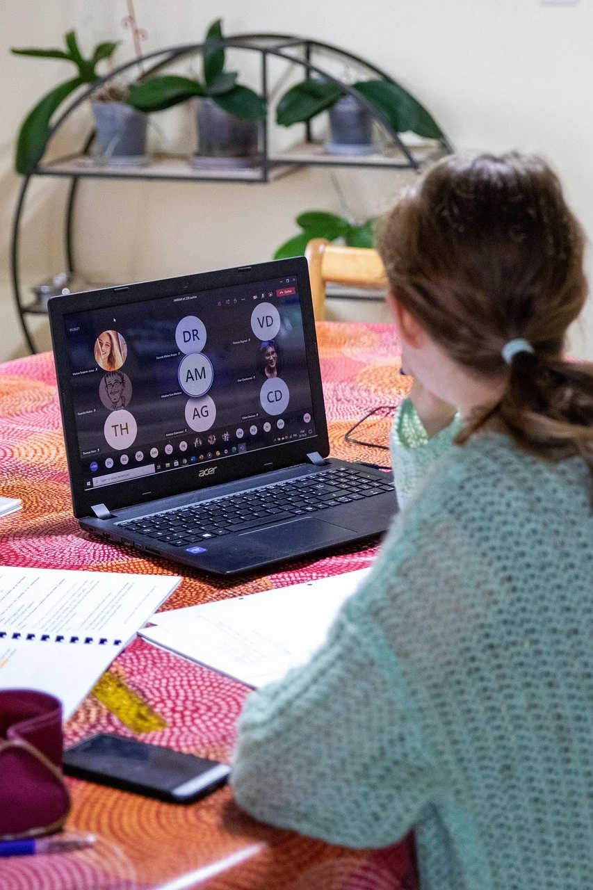 A girl with brown hair in a ponytail working on a laptop during a virtual meeting, with papers and a smartphone on a colorful tablecloth.