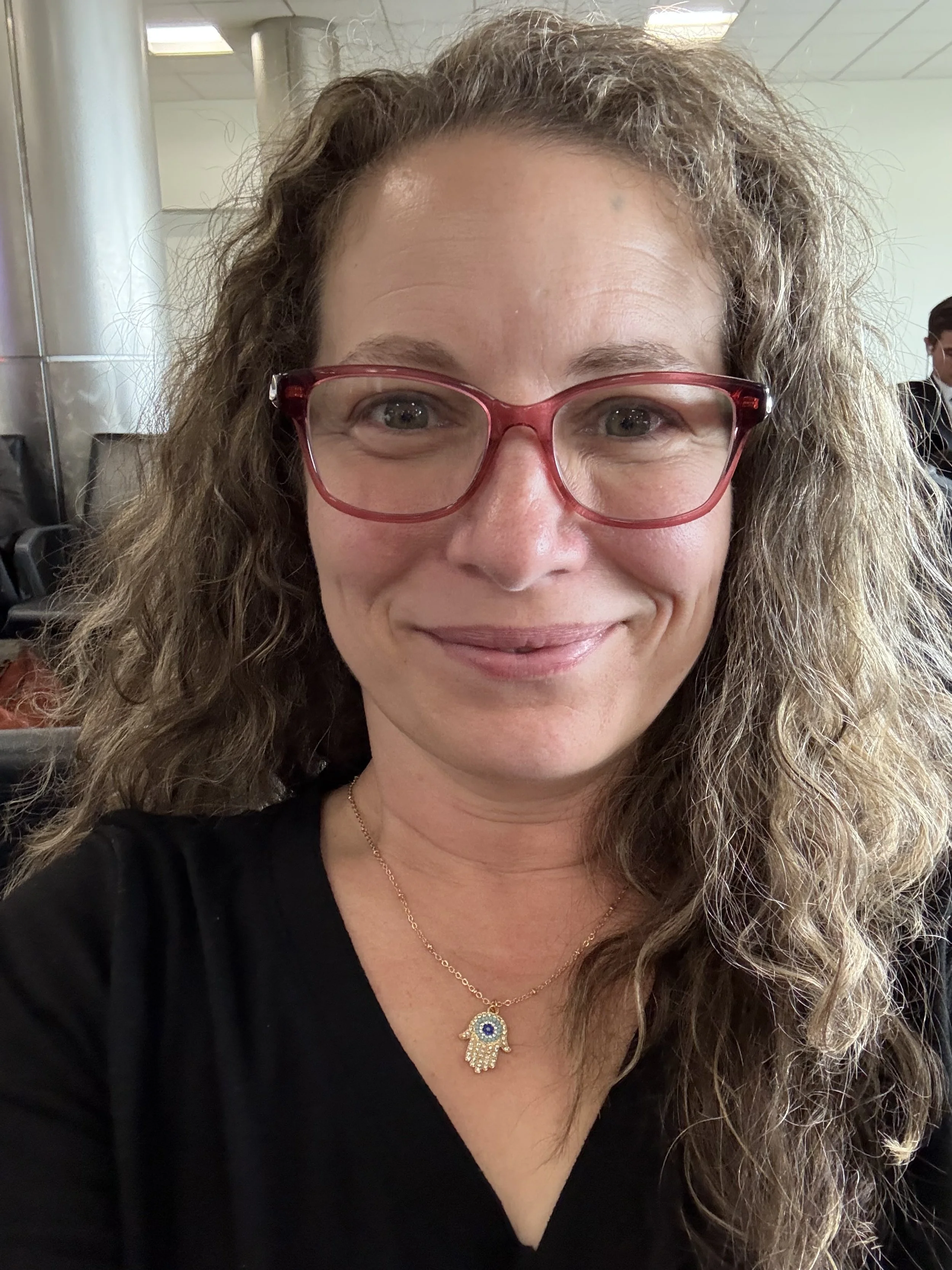 A woman with curly brown hair and light skin wearing red glasses, a black top, and a gold necklace with a Hamsa hand pendant. She is smiling and seated indoors.
