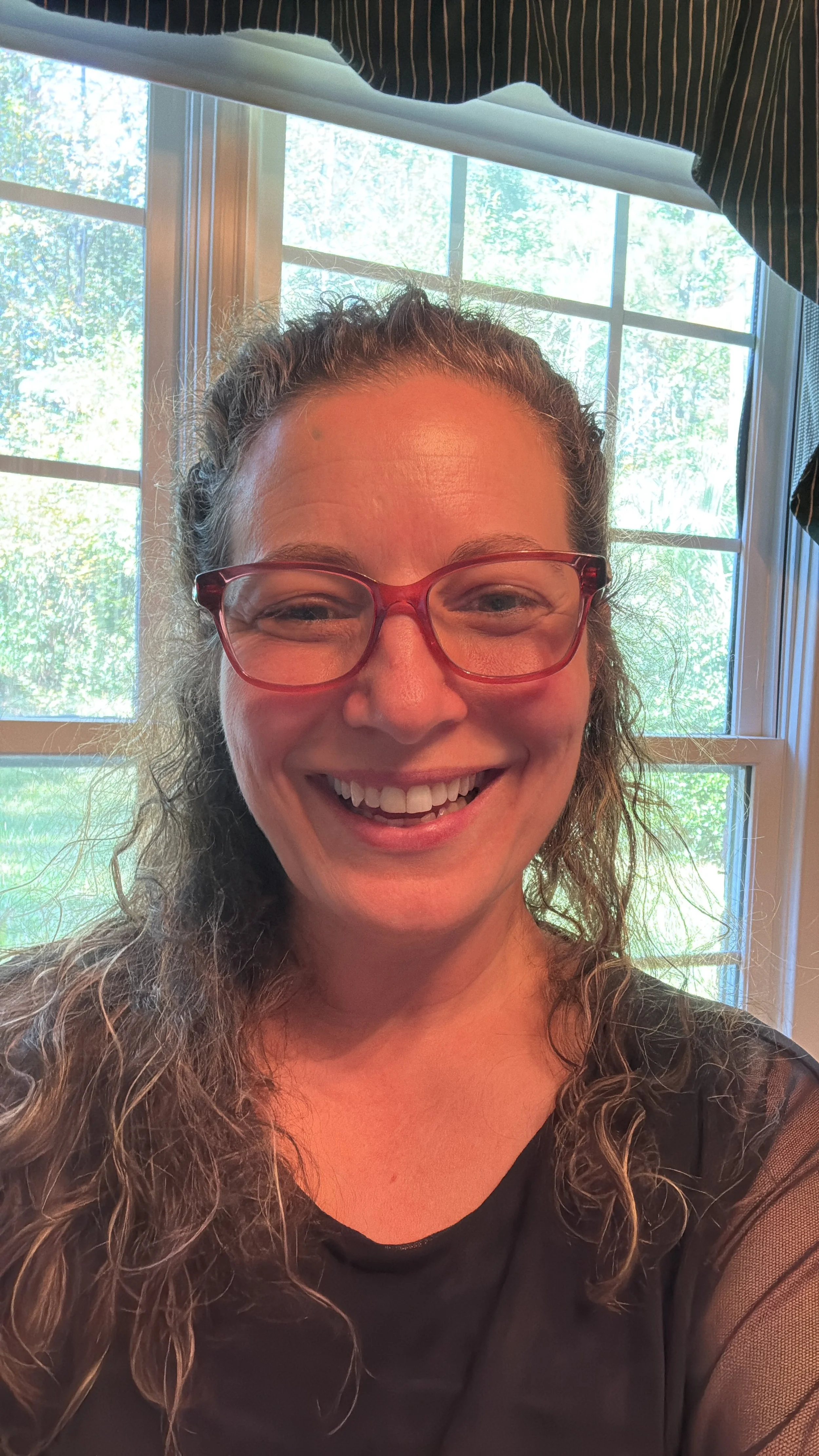 A woman with curly hair and red glasses smiling at the camera with a window and green trees visible behind her.