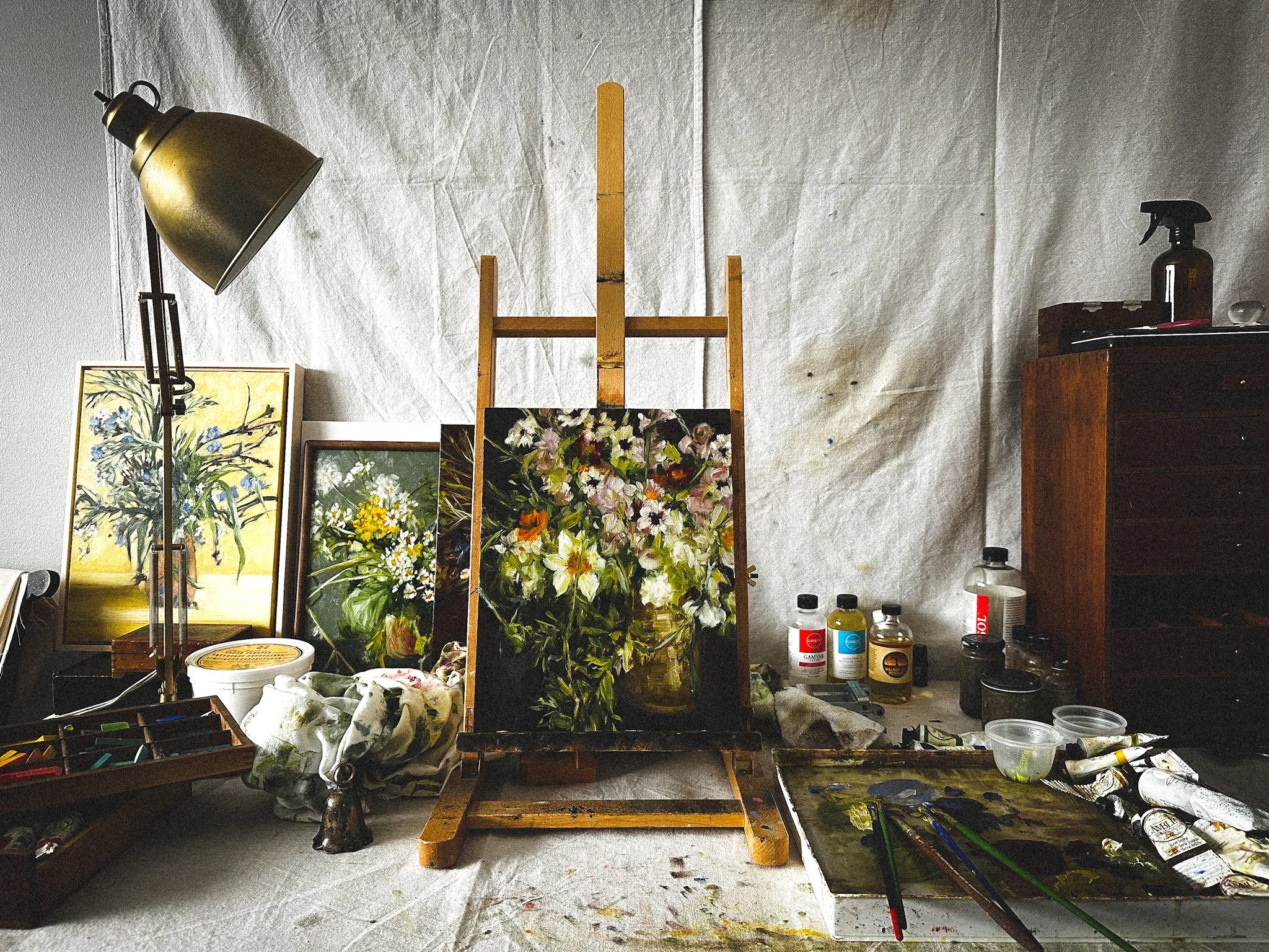 An artist's workspace with two floral paintings, paint supplies, and a wooden easel on a white tarpaulin background.