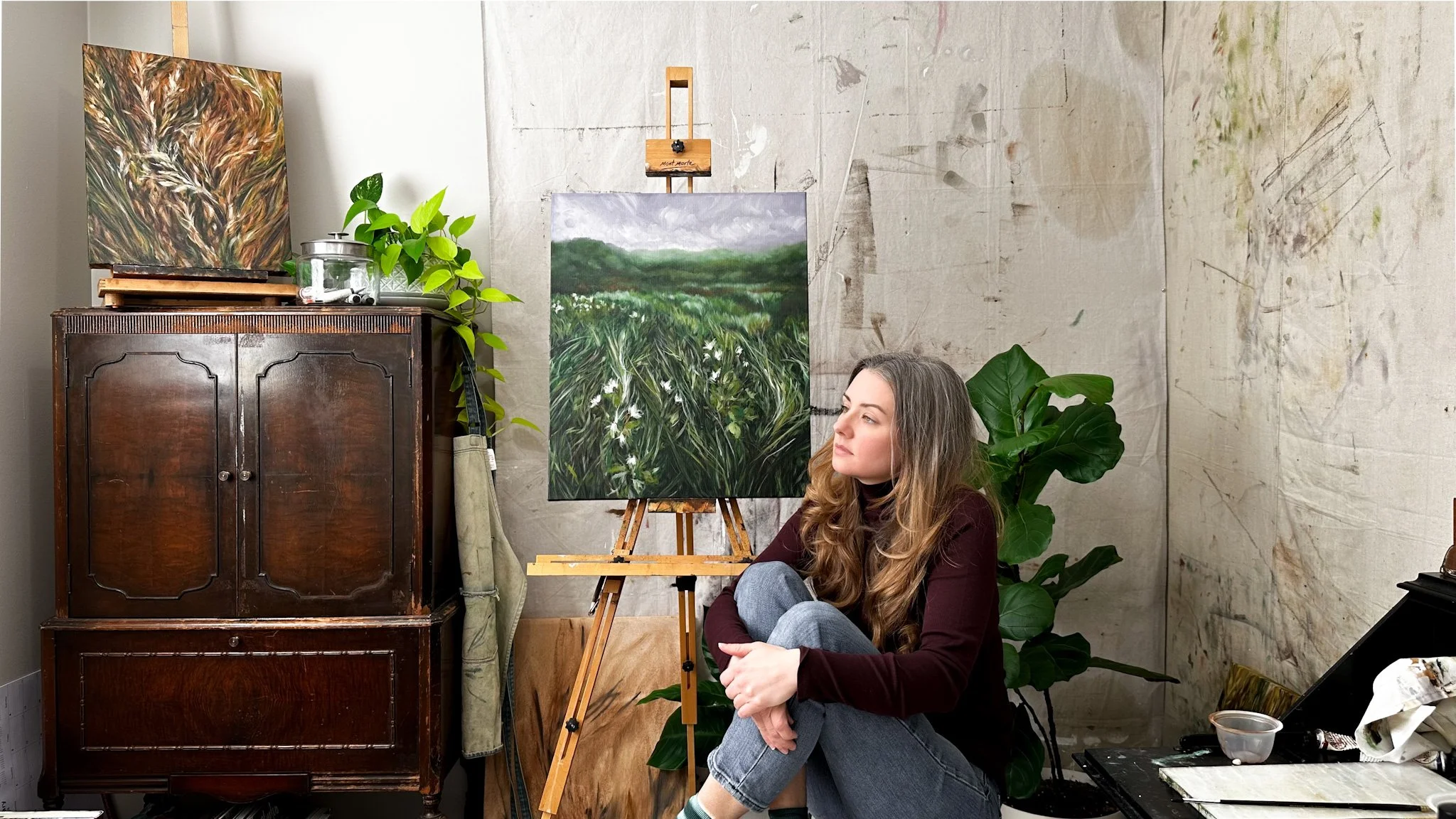 A woman with long hair sitting on the floor of an art studio next to a painting on an easel and a wooden cabinet with another painting on top. There are various art supplies and a large green plant nearby.