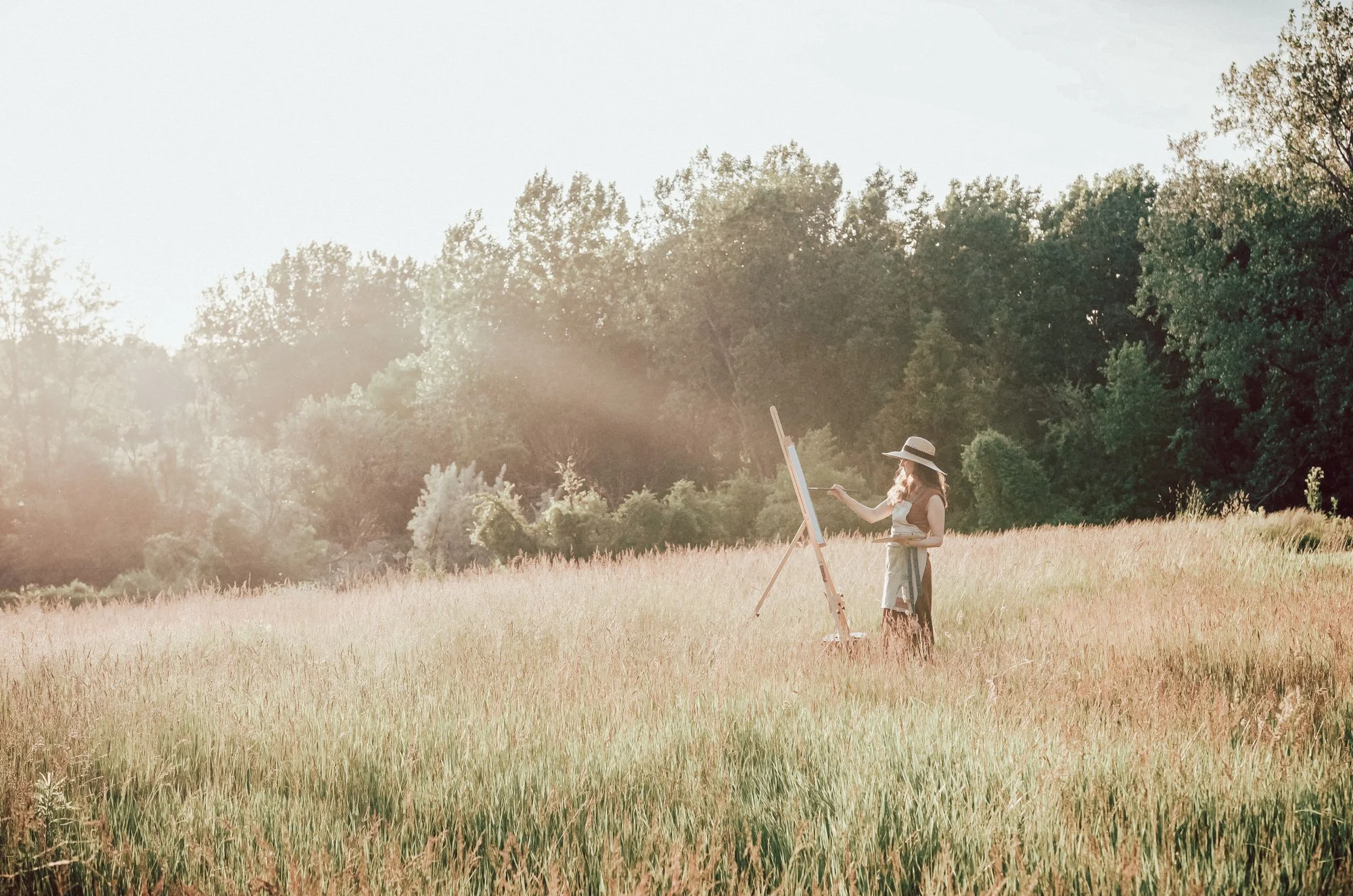 A woman painting outdoors on a sunny day in a field near trees, wearing a sun hat and dress.