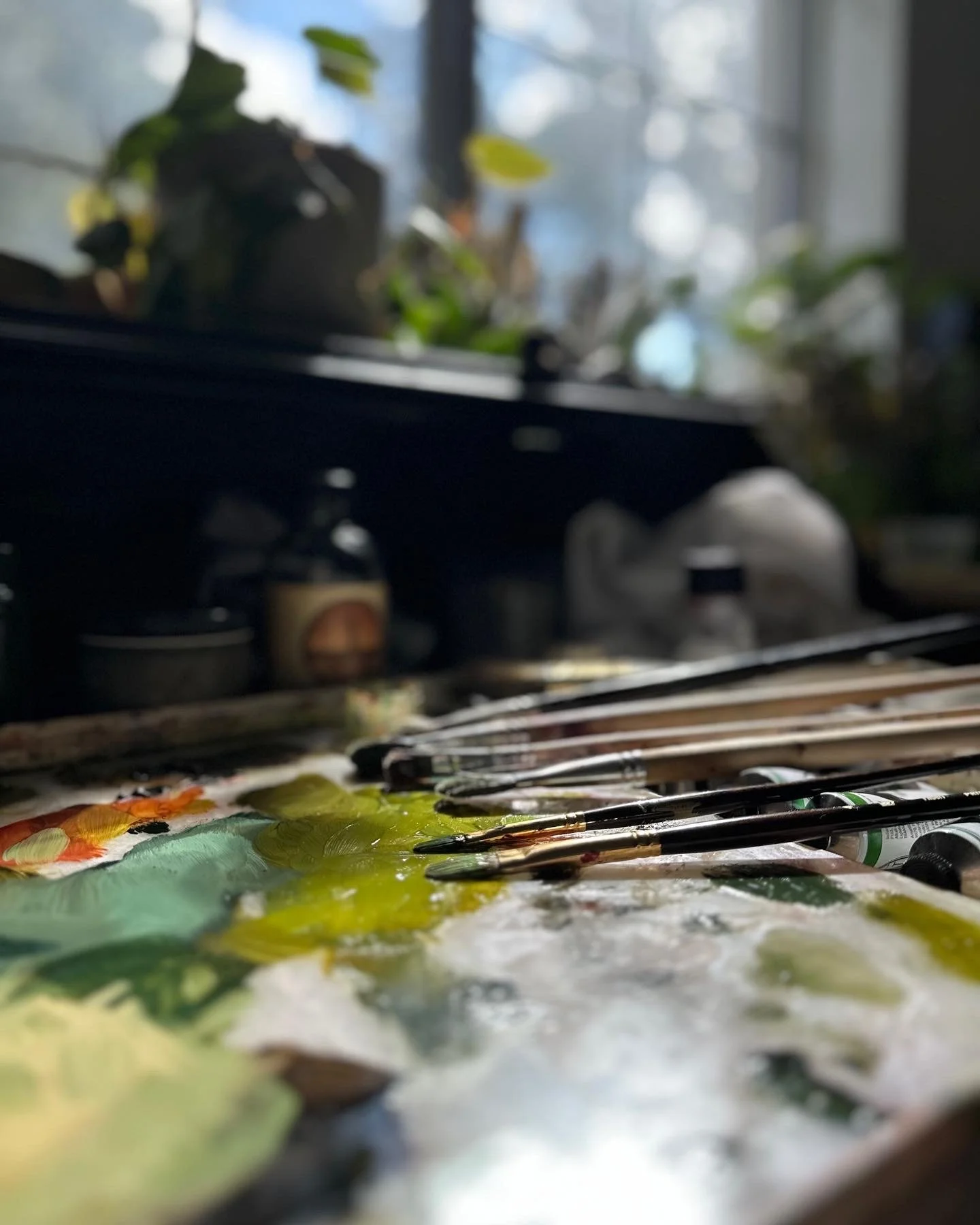 Paintbrushes and tubes of paint on a palette in front of a window with plants.