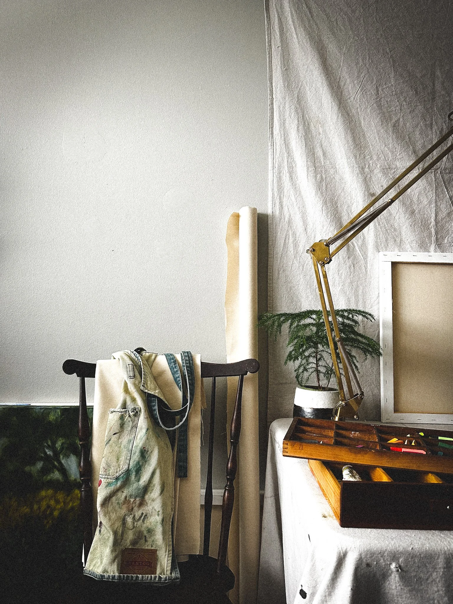 Interior scene with a chair, painting, apron, plant, lamp, and a table with art supplies.