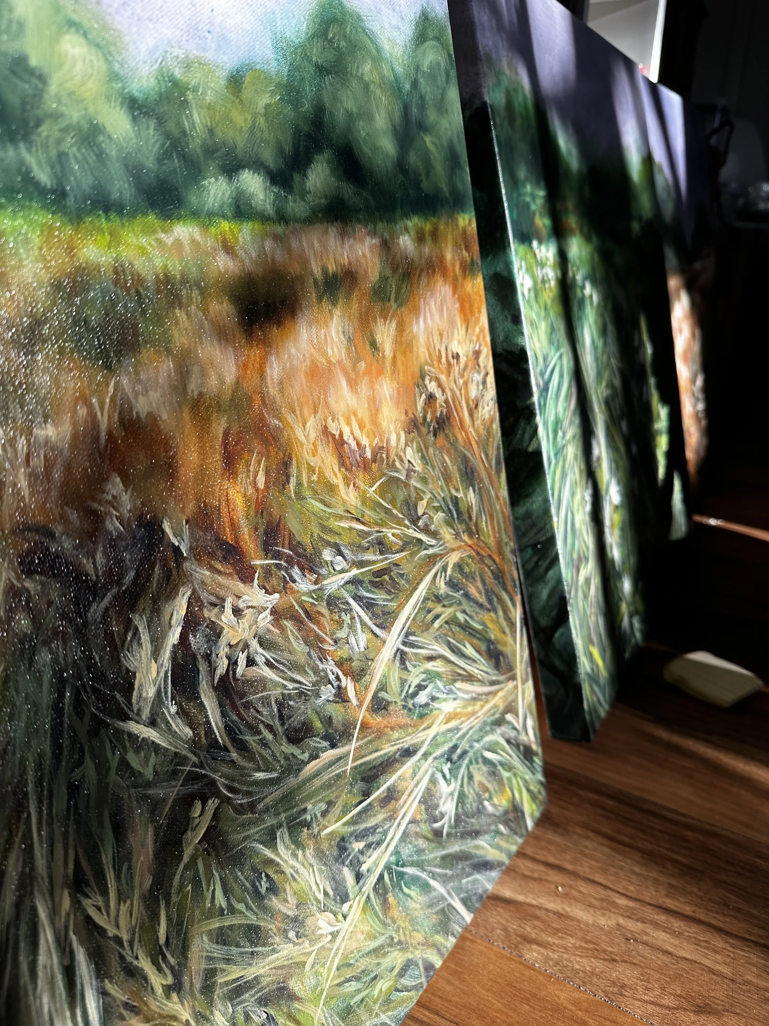 Close-up view of multiple landscape paintings with green and earthy tones, leaning against a wall on a wooden floor.