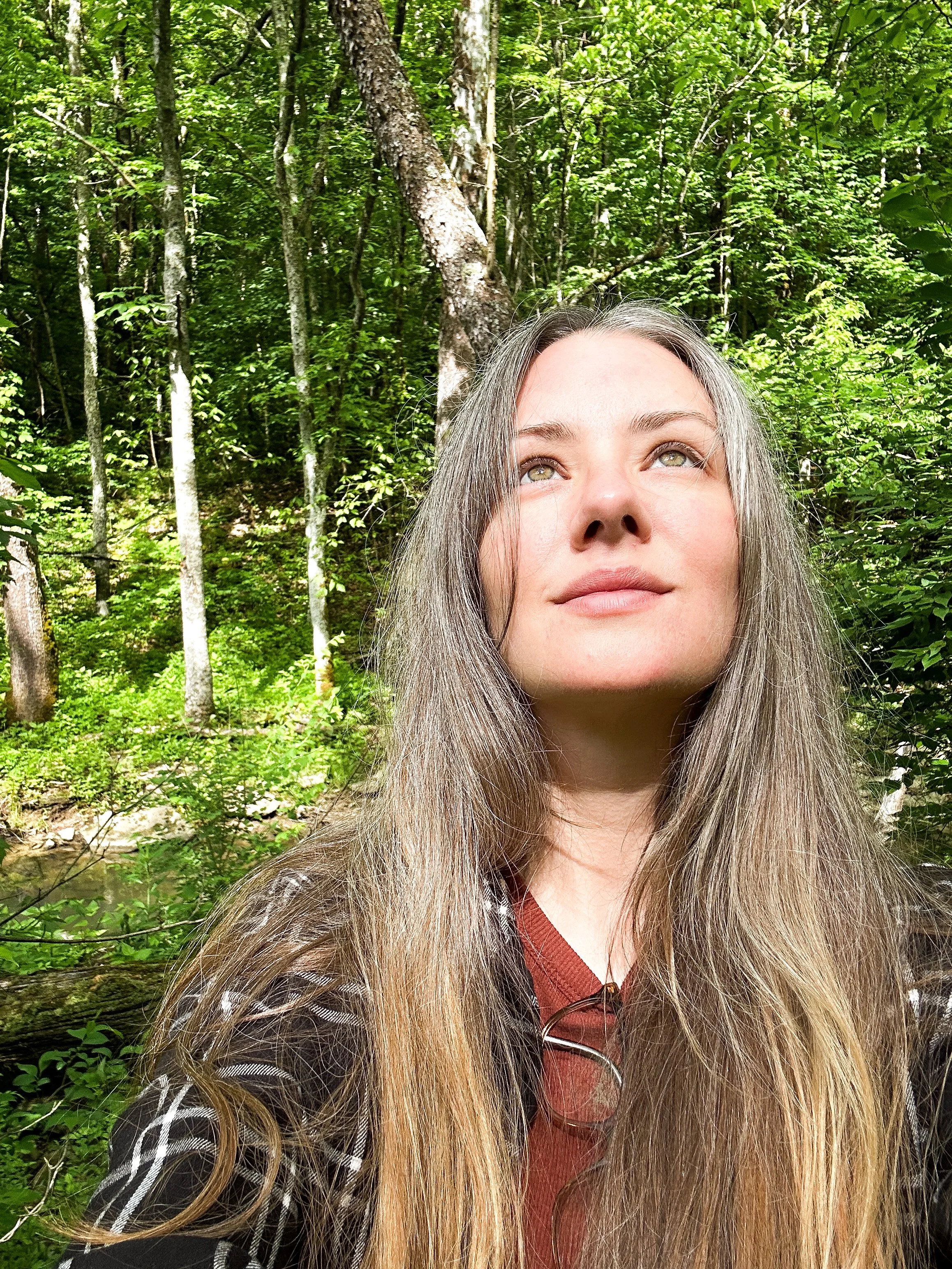 A woman with long, grayish hair looking upward in a lush green forest.