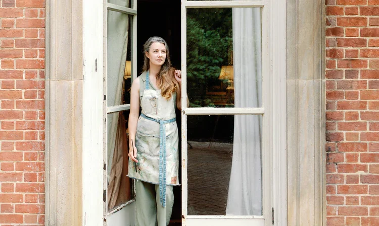 A woman standing in an open window of a brick building, looking outside with a thoughtful expression.