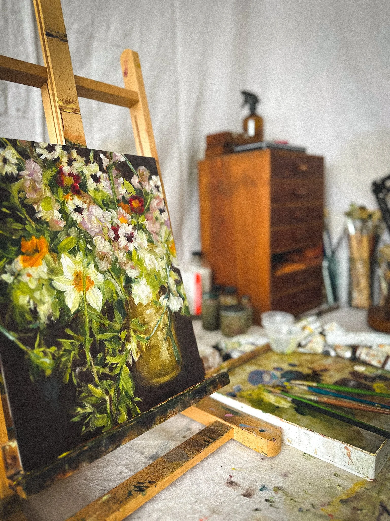 An easel holding a small, colorful floral still life painting in an artist's studio. The workspace includes brushes, paint tubes, and art supplies on a table, with a wooden dresser and art equipment in the background.