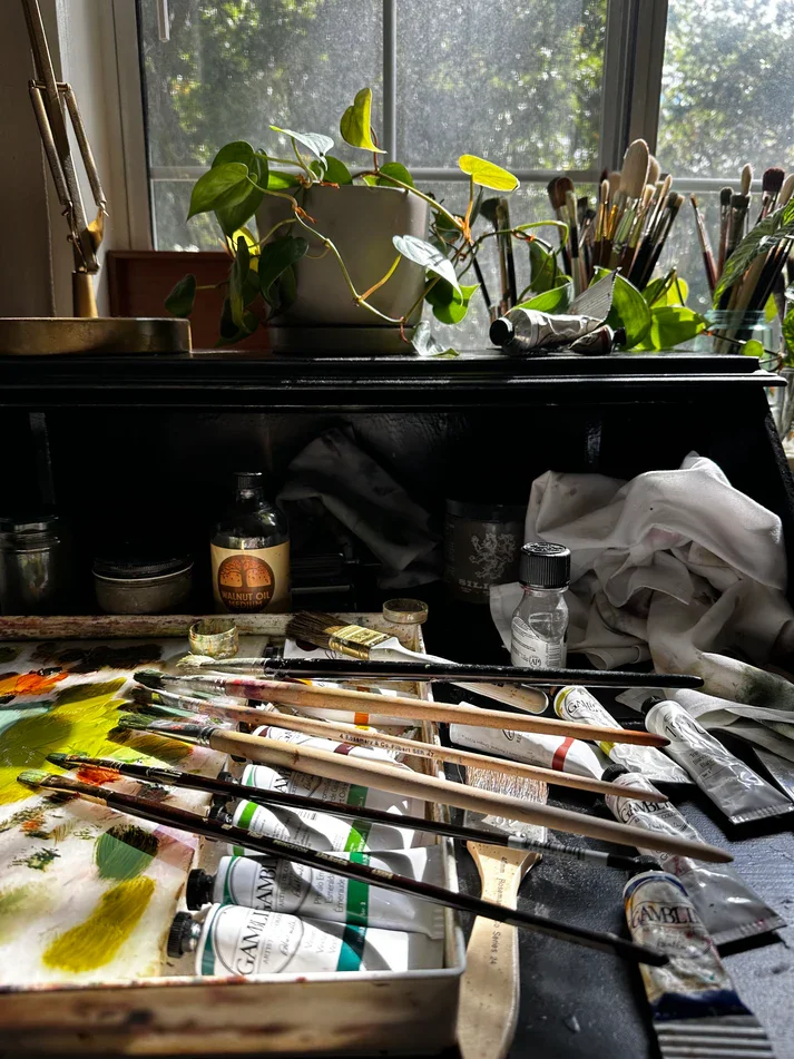An artist's workspace with paintbrushes, paint tubes, and a palette on a cluttered table near a window with sunlight and green trees outside. A potted plant is on the windowsill.