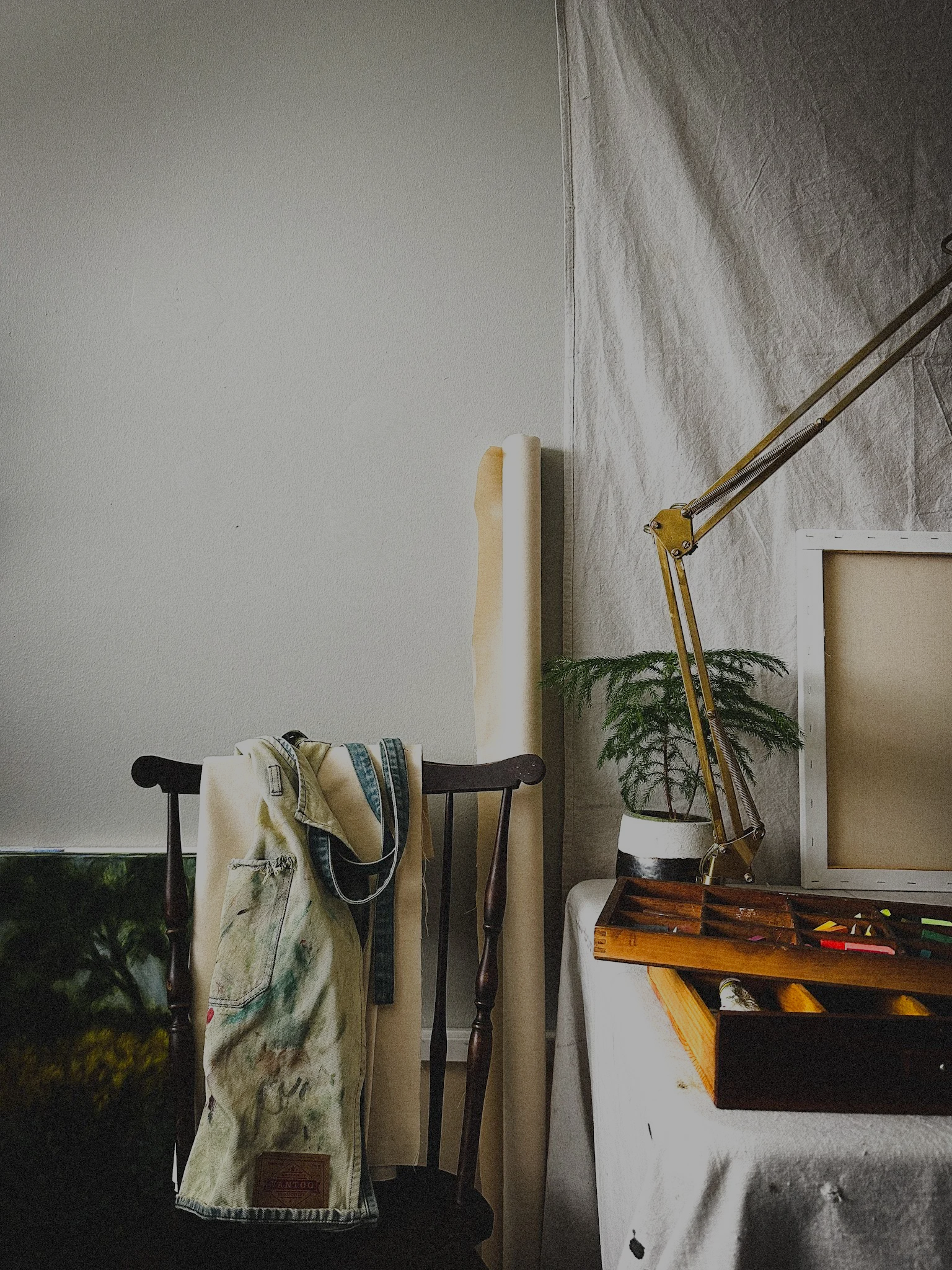 An artist's workspace with a painting of colorful flowers on an easel, various art supplies, and paintings leaning against the wall, illuminated by a large adjustable lamp.