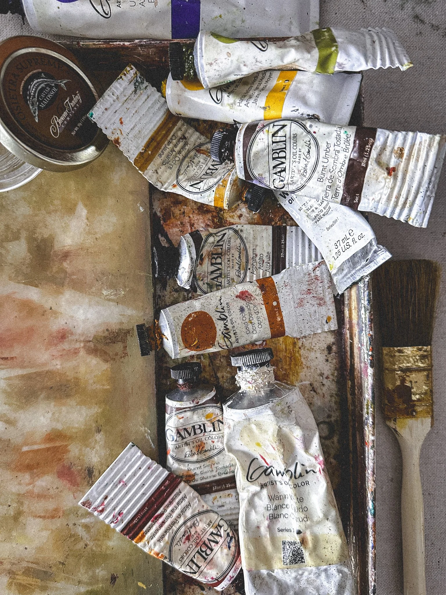 Assorted paint tubes, palette knife, and containers on a dirty artist's palette, with some paint residue and smudges.