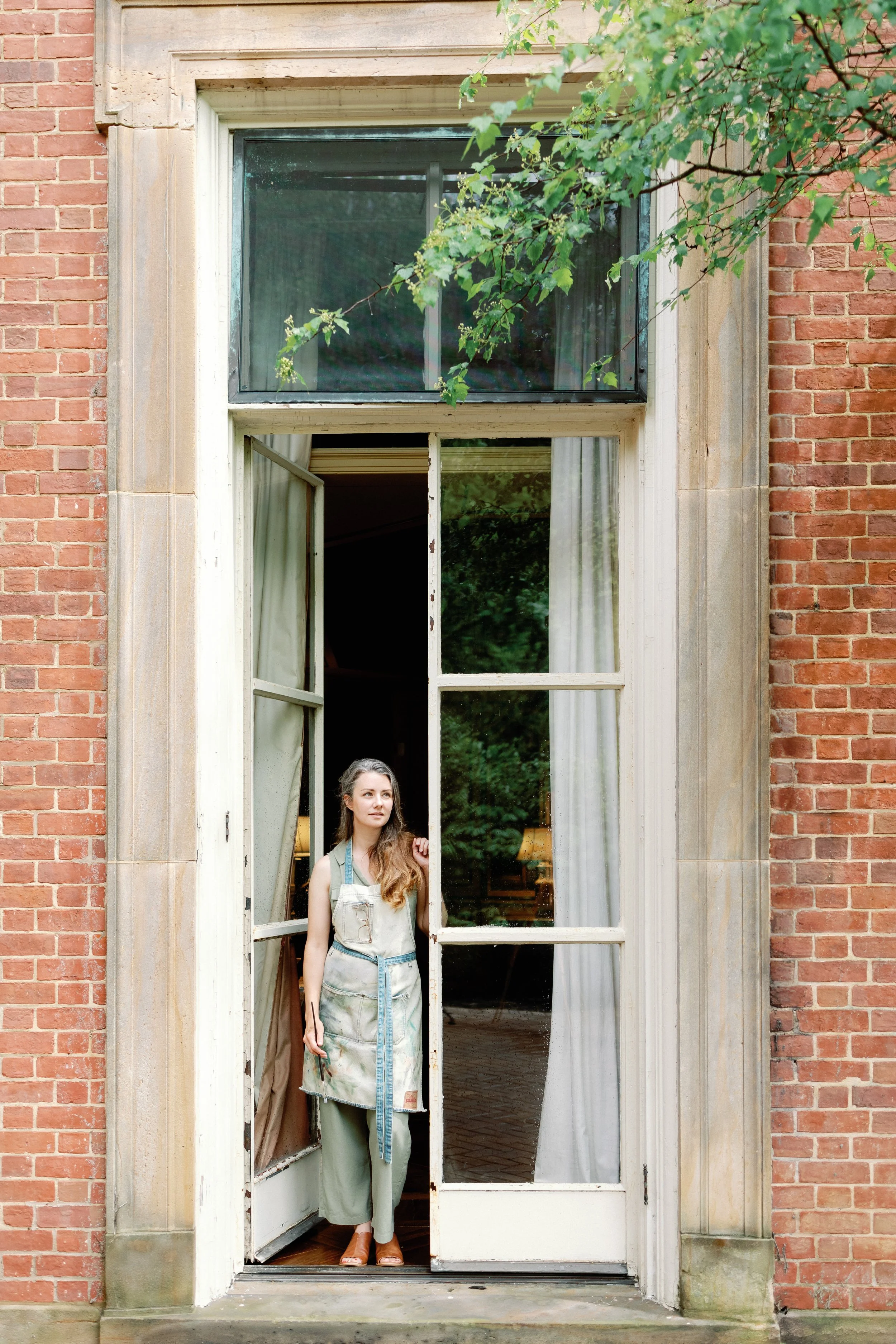 A woman standing behind a large old window, looking outside, with brick walls on either side and a tree branch with green leaves above.