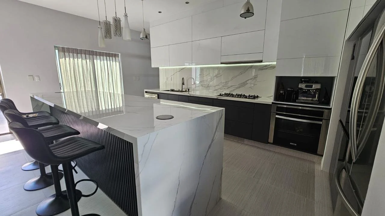 Cocina moderna con encimera y paredes de mármol blanco, electrodomésticos de acero inoxidable y barra con sillas negras.