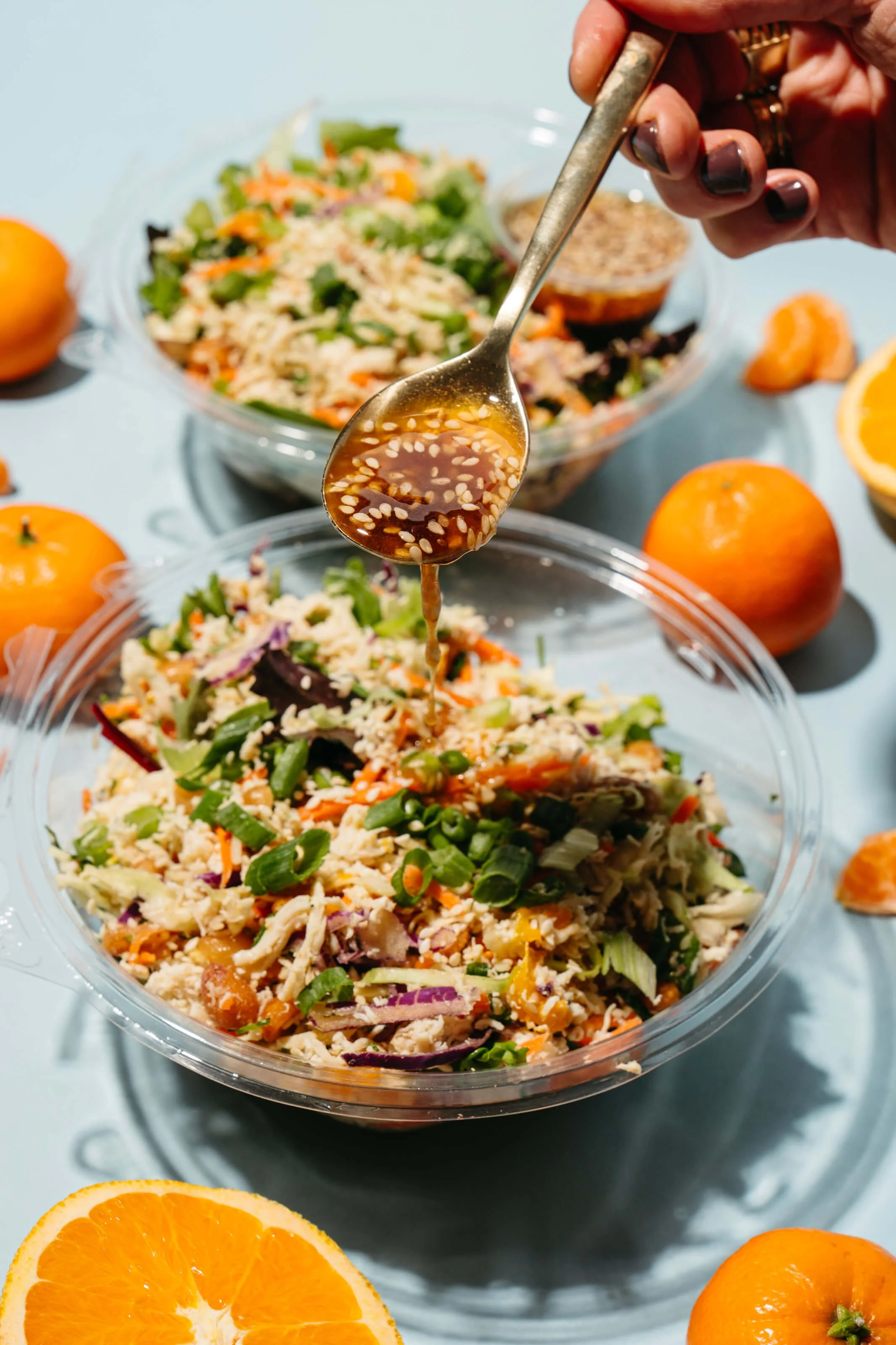 A hand drizzling salad dressing over a bowl of mixed vegetable salad with chopped green onions, carrots, purple cabbage, and shredded chicken, surrounded by whole oranges and halved orange slices on a light blue surface.