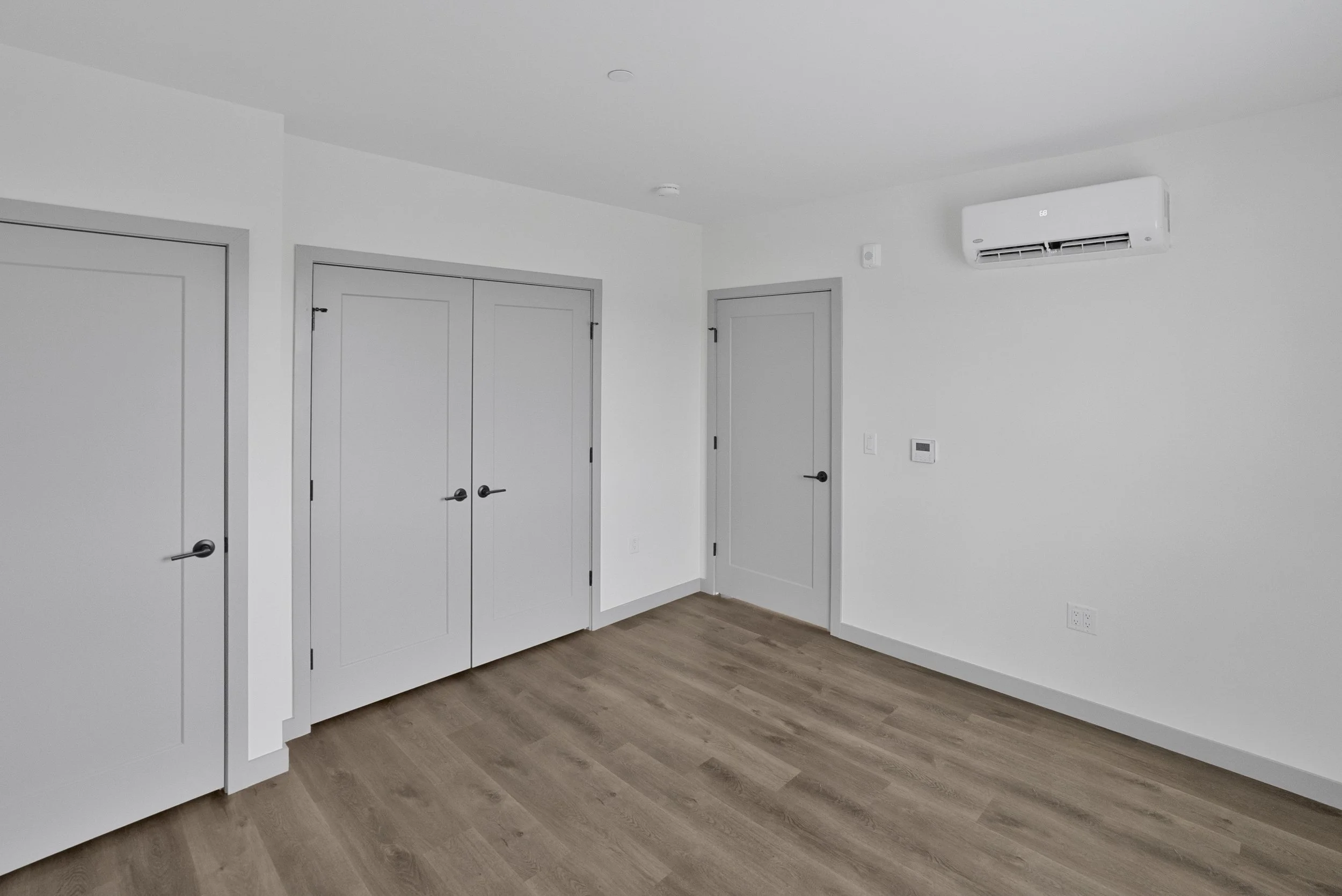 A corner of a room with three white doors, gray door frames, a white wall-mounted air conditioning unit, a small thermostat, and wood flooring.