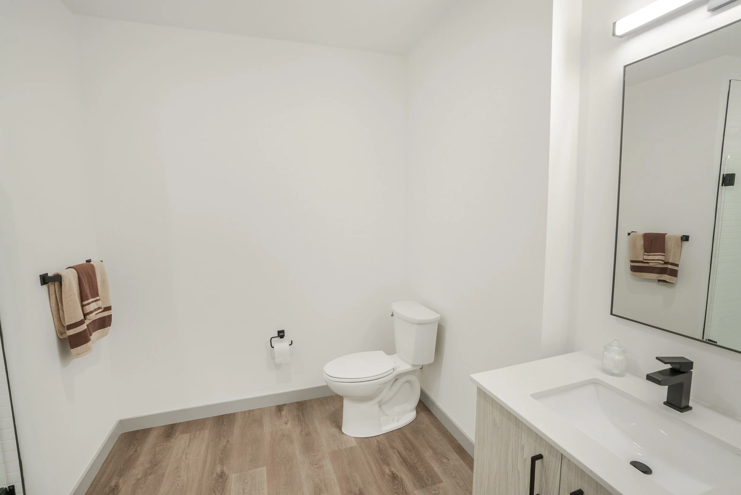A modern, minimalistic bathroom with a white toilet, a mirror above the sink, a black faucet, and beige towels hanging on black towel racks.