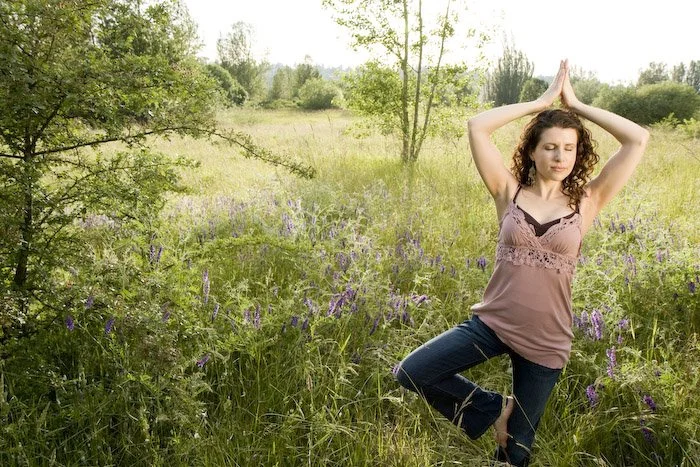 A woman practicing yoga outdoors in a grassy field with purple wildflowers, standing in a tree pose with hands pressed together above her head and eyes closed.