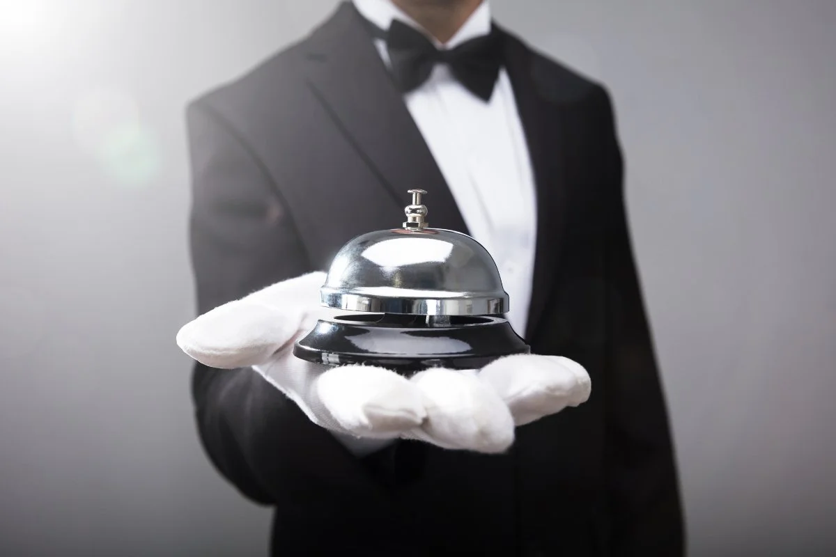 A concierge dressed in a black tuxedo with a bow tie holding a hotel desk bell with a white glove.