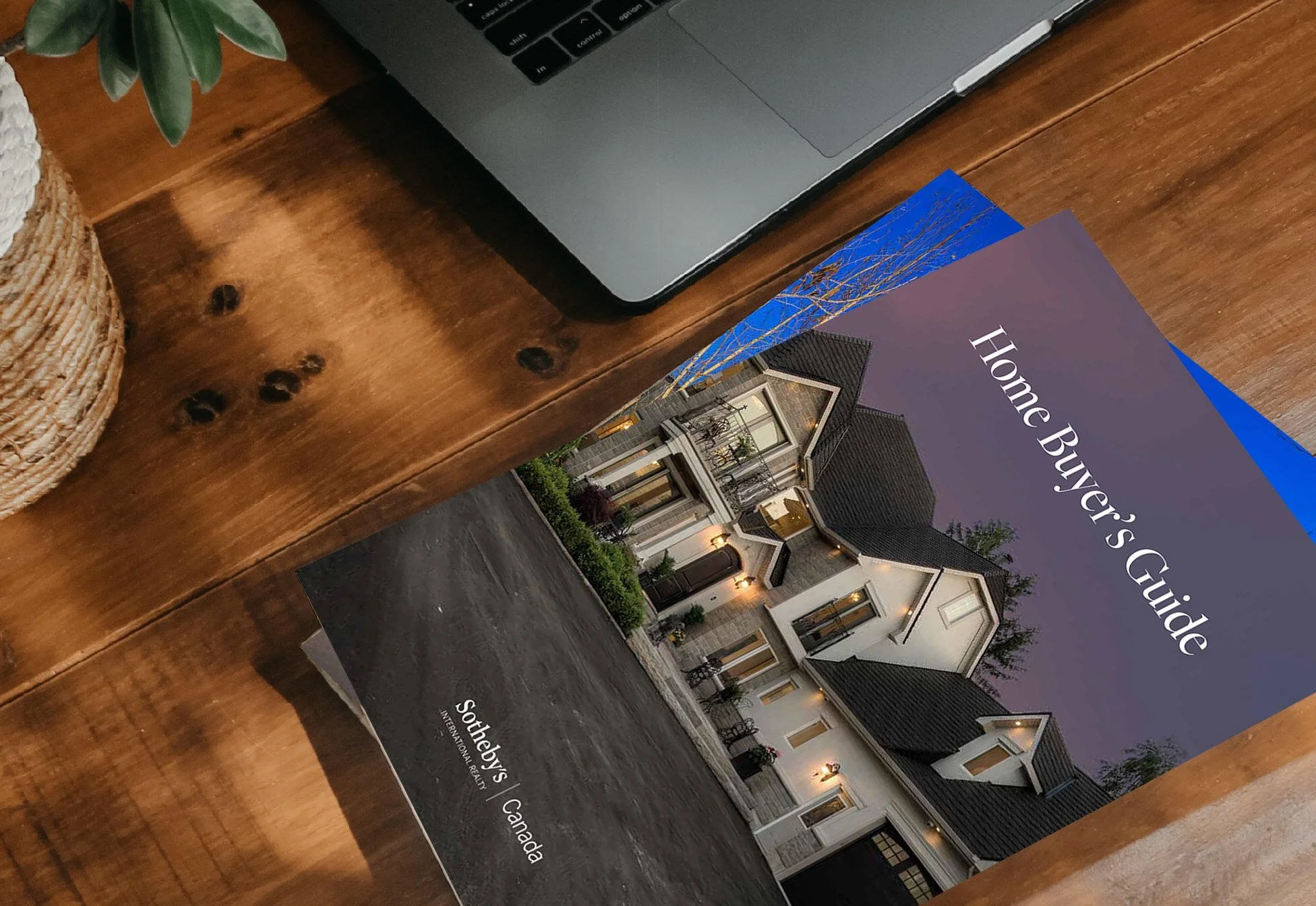 Book titled 'Home Buyer’s Guide' lying on a wooden desk next to a laptop, with part of a plant and a woven container nearby.