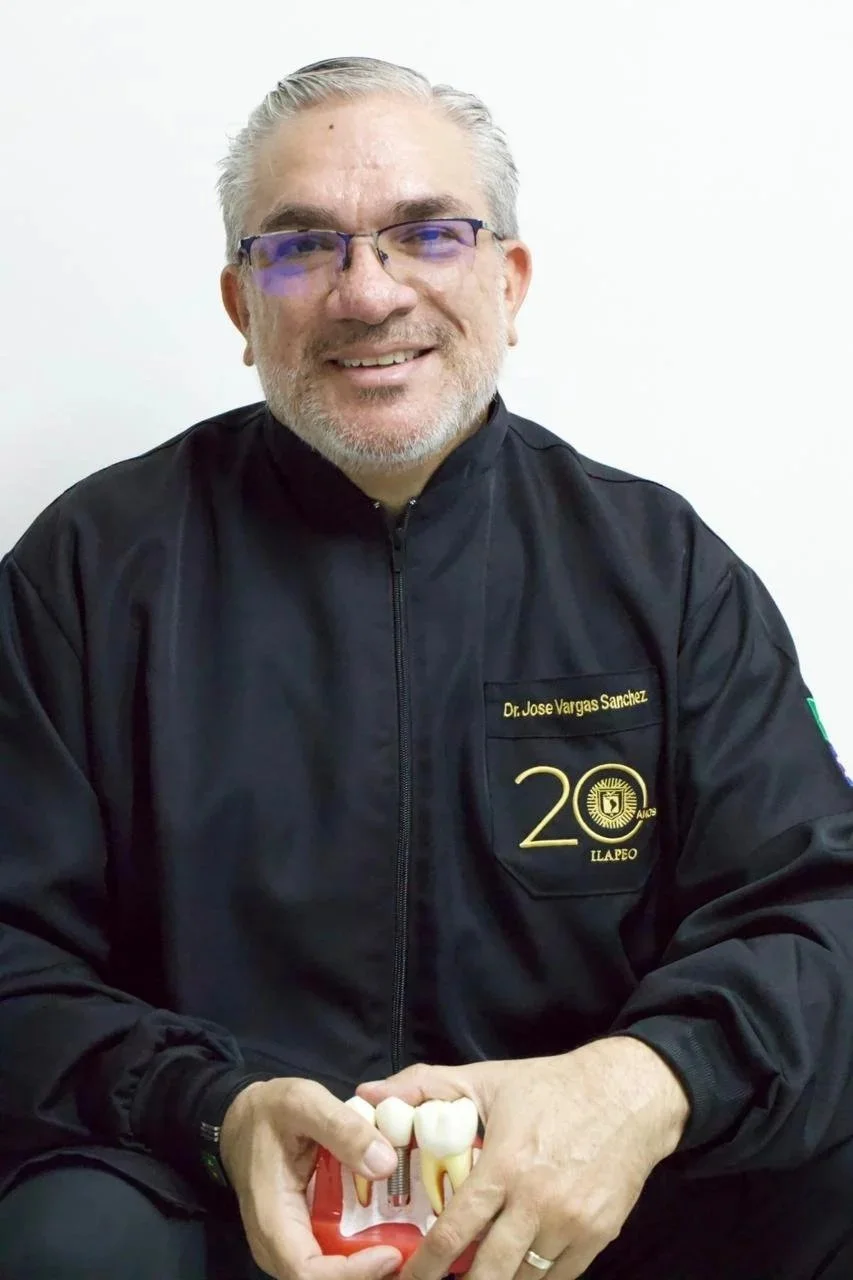 Hombre con gafas y barba, vestido con chaqueta negra con un logotipo del 20 aniversario en la camiseta, sosteniendo un modelo de dientes y implantes dentales.
