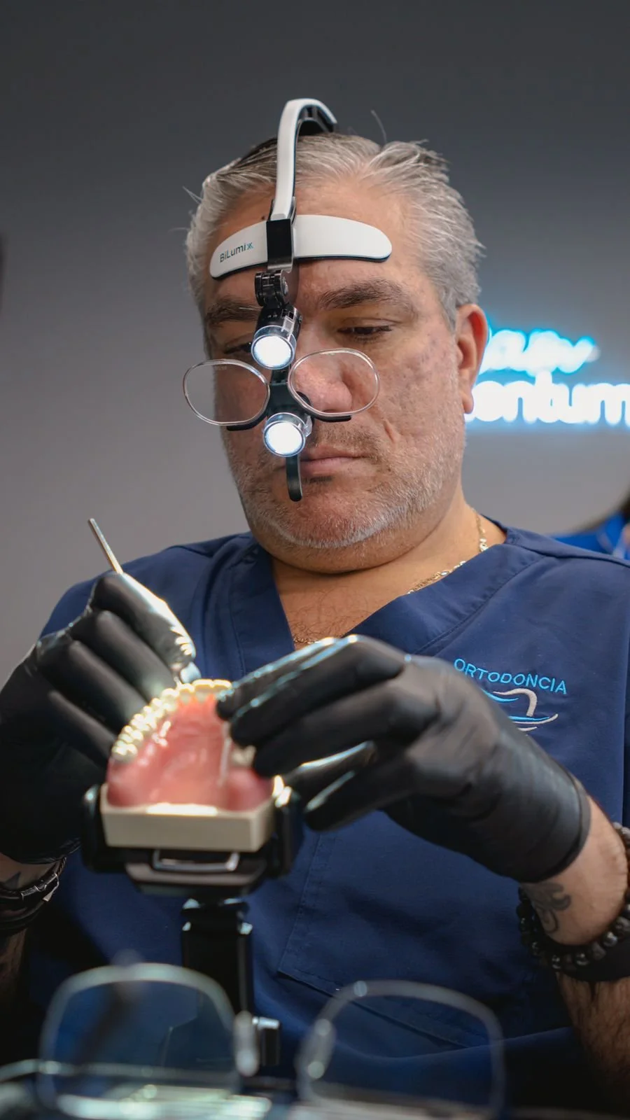 Dentista en consulta usando lupas y luz especial, trabajando en un modelo dental con instrumentos.
