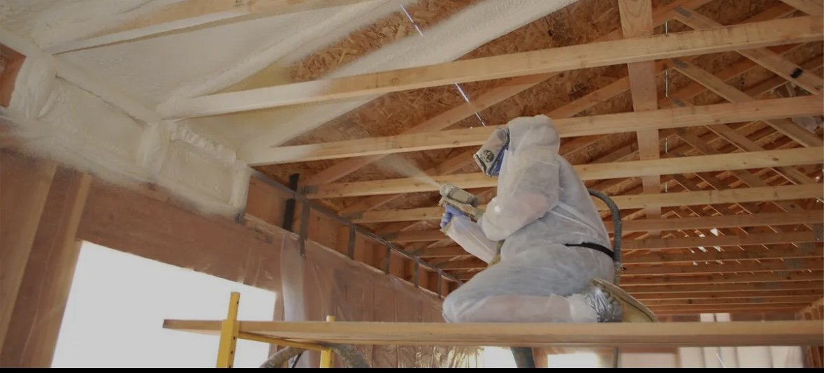 A construction worker dressed in protective gear working on the ceiling framework of a building, kneeling on a ladder and installing or inspecting wooden roof trusses.