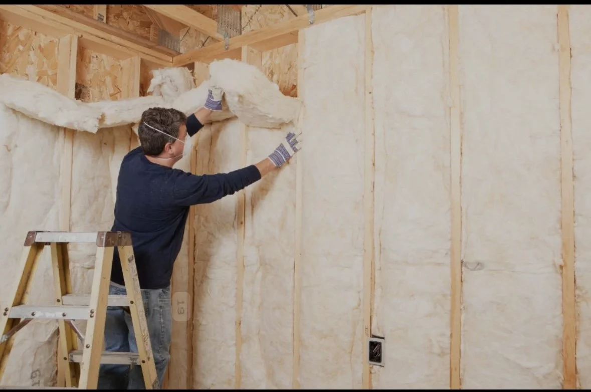 A person installing insulation in an unfinished wall of a building, wearing a mask and gloves, standing on a small wooden ladder.