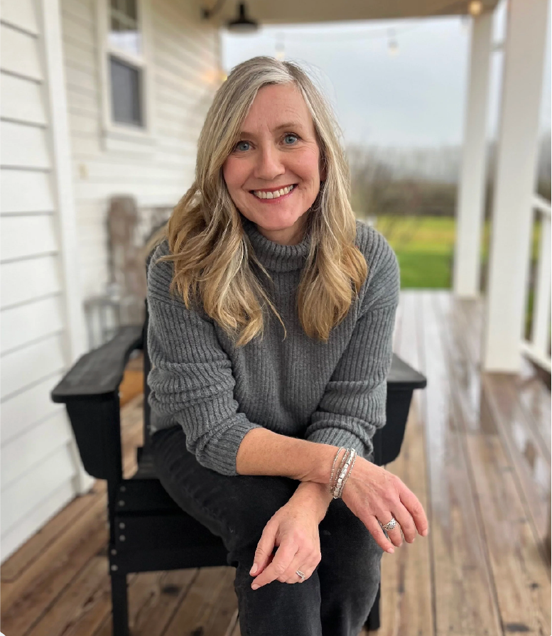 A woman with blonde hair and blue eyes smiling, sitting outdoors on a porch with white siding, wearing a gray turtleneck sweater and black pants, with a landscape of green grass and trees in the background.