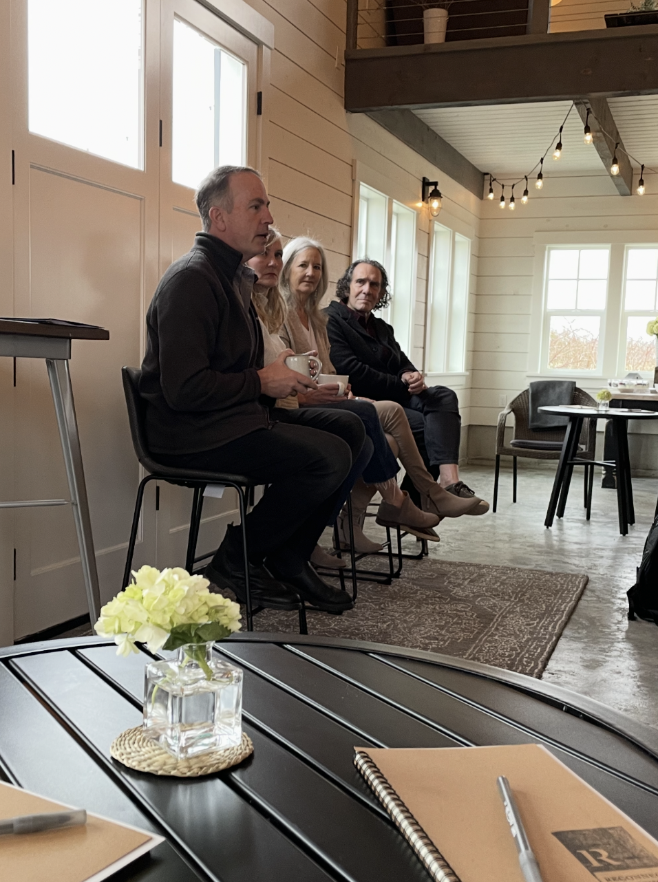 Four people sitting on chairs in a rustic, well-lit room, engaged in a discussion, with a table in the foreground holding a small vase with white flowers, notebooks, and a pen.