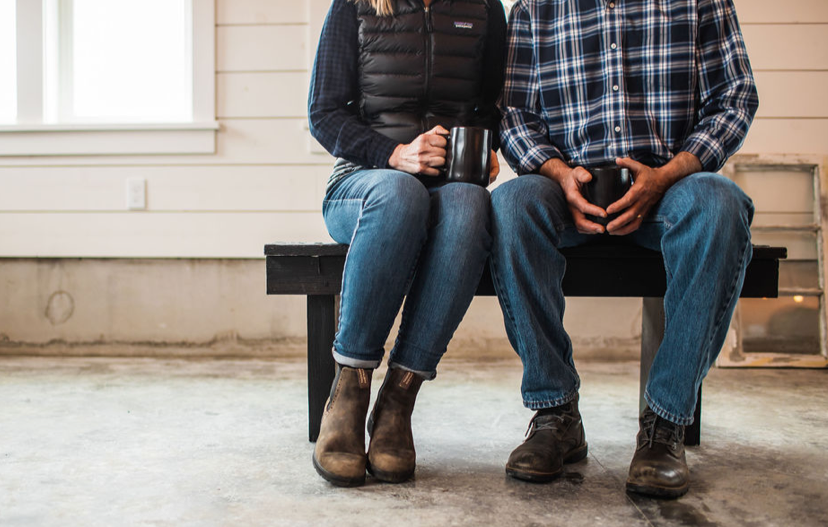 Two people sitting on a black bench, holding mugs, in a room with white walls and concrete floor.