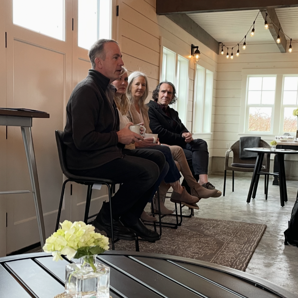 Four people seated on stools in a cozy room with large windows and string lights, having a discussion or presentation, with a small table with a flower vase in the foreground.