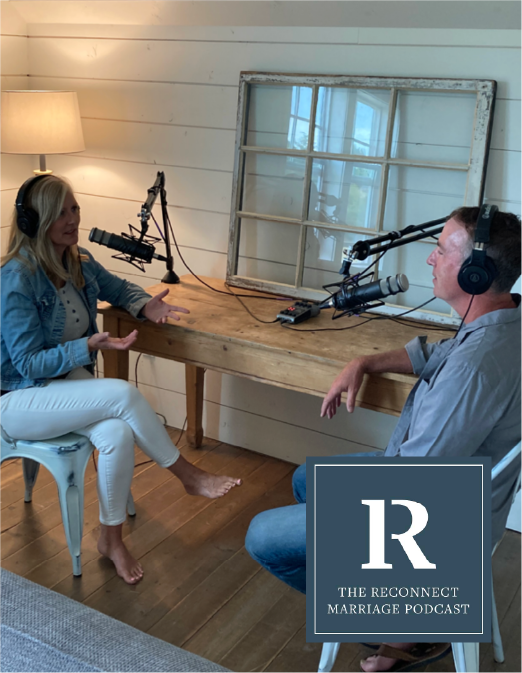 Two people are recording a podcast in a cozy, rustic room with white wooden walls. The woman on the left is wearing a denim jacket and white pants, sitting barefoot, speaking into a microphone with headphones on. The man on the right is wearing a gray shirt and blue jeans, speaking into a microphone with headphones. A large mirror leans against the wall behind them, and a window reflects natural light into the room. A lamp is on, creating warm lighting. The podcast is titled 'The Reconnect Marriage Podcast'.