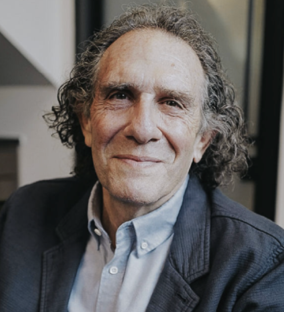 A middle-aged man with gray curly hair, smiling, wearing a light blue collared shirt and a dark jacket.