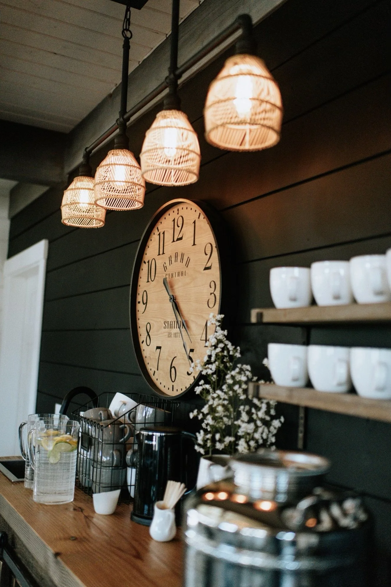 Coffee station with a wooden countertop, a glass pitcher of lemon water, white mugs, a black coffee maker, and a small vase with white flowers. A large wooden clock on dark wall, and warm overhead lighting.