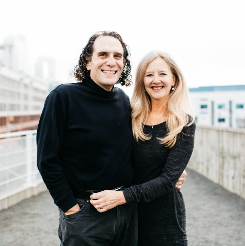 A smiling man and woman standing on a rooftop with city buildings in the background, hugging and looking at the camera.