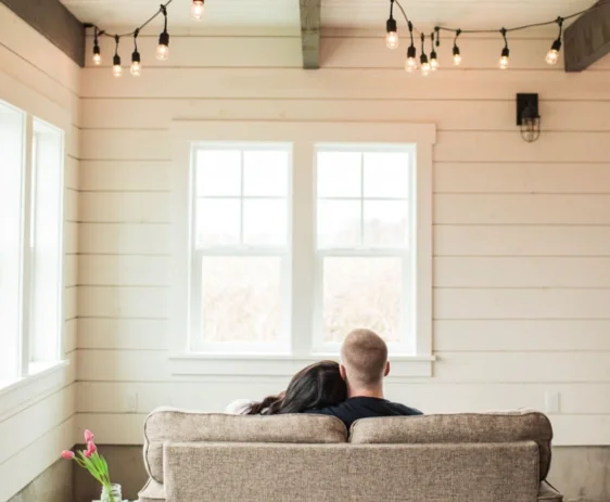 A couple sitting on a beige couch in front of windows in a cozy, well-lit room with string lights hanging from the ceiling.