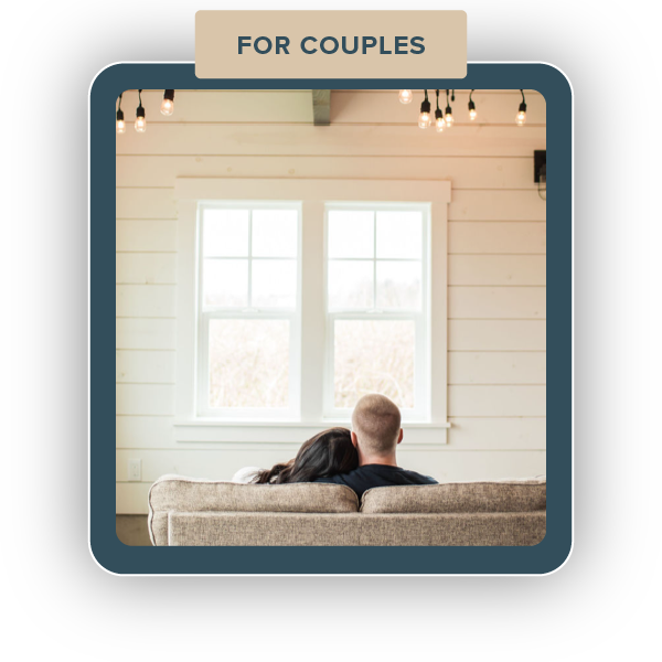 A couple sitting on a couch, facing a window in a cozy, white-walled room.