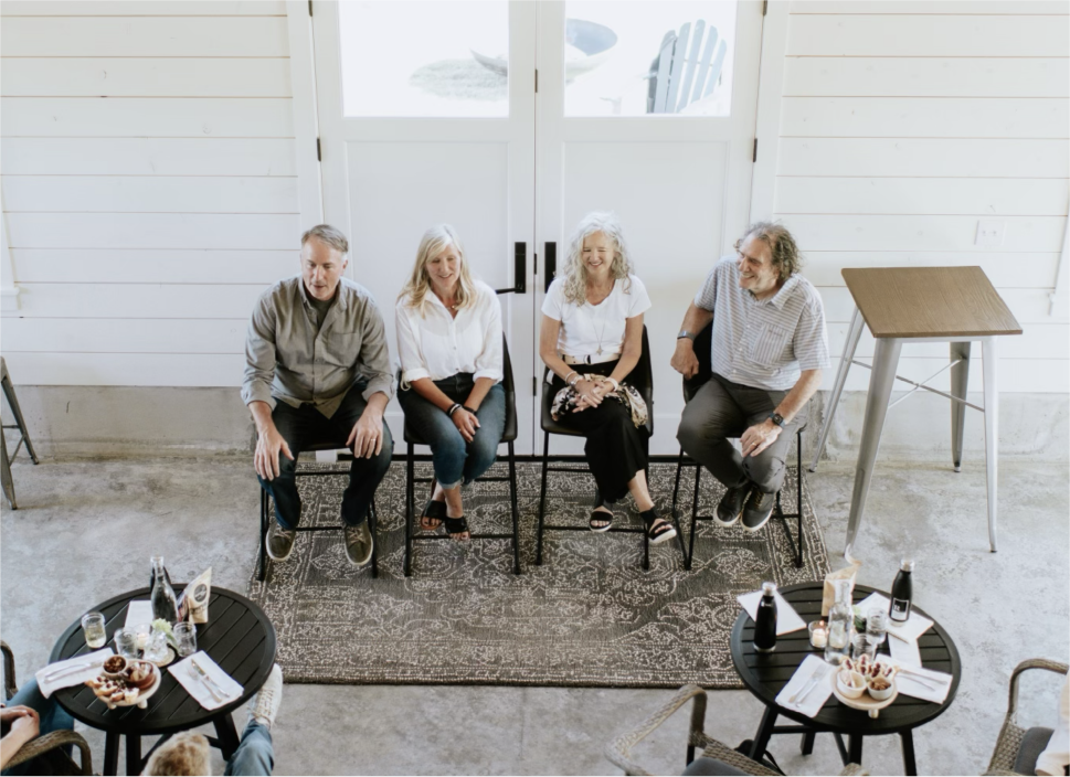 Four older adults sitting on chairs on a rug in a white room, with tables set with snacks and drinks nearby.