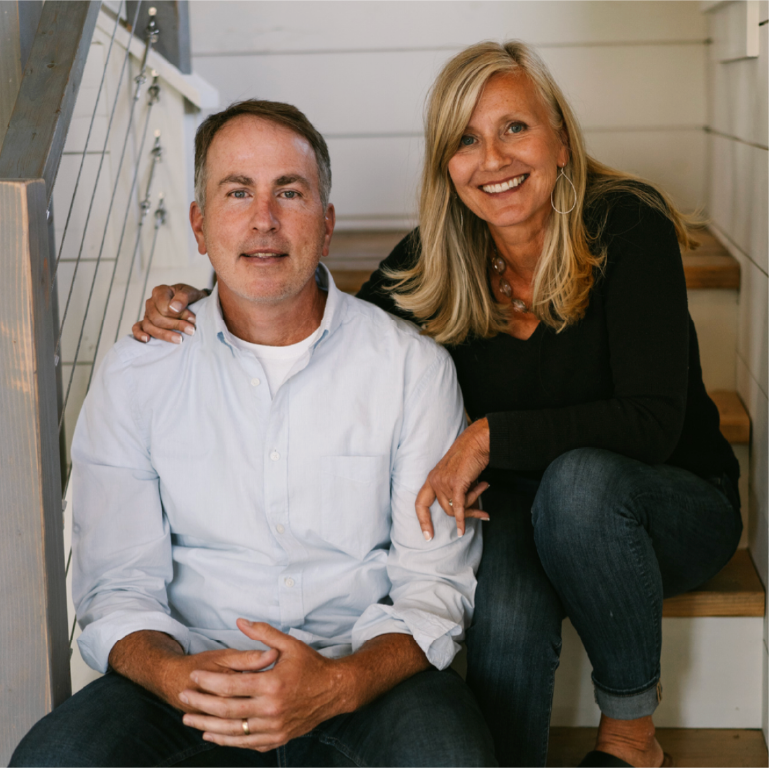 A smiling middle-aged man and woman sitting on wooden stairs inside a house. The man wears a white button-up shirt, and the woman wears a black top with hoop earrings, and has her arm around the man's shoulder.