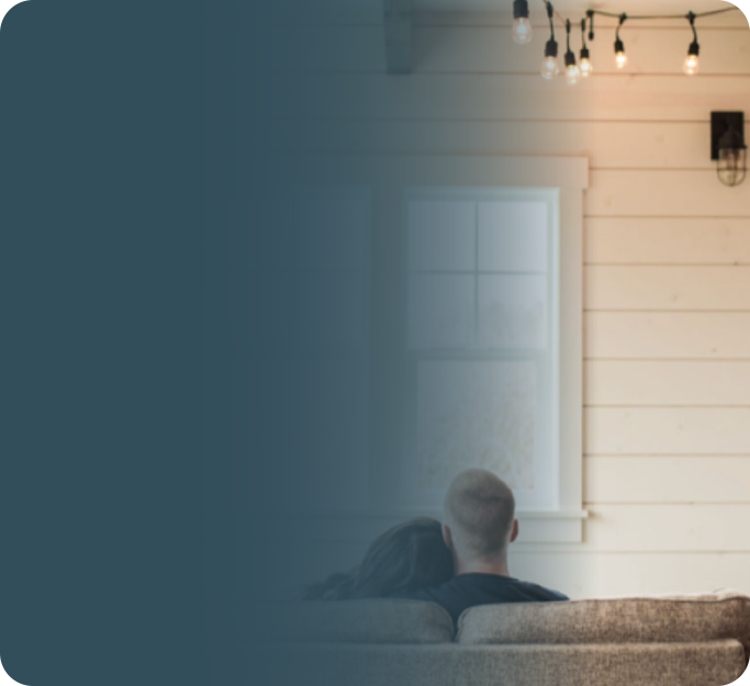 A couple sitting on a beige couch, watching a small television inside a cozy room with white wooden walls and string lights hanging from the ceiling.