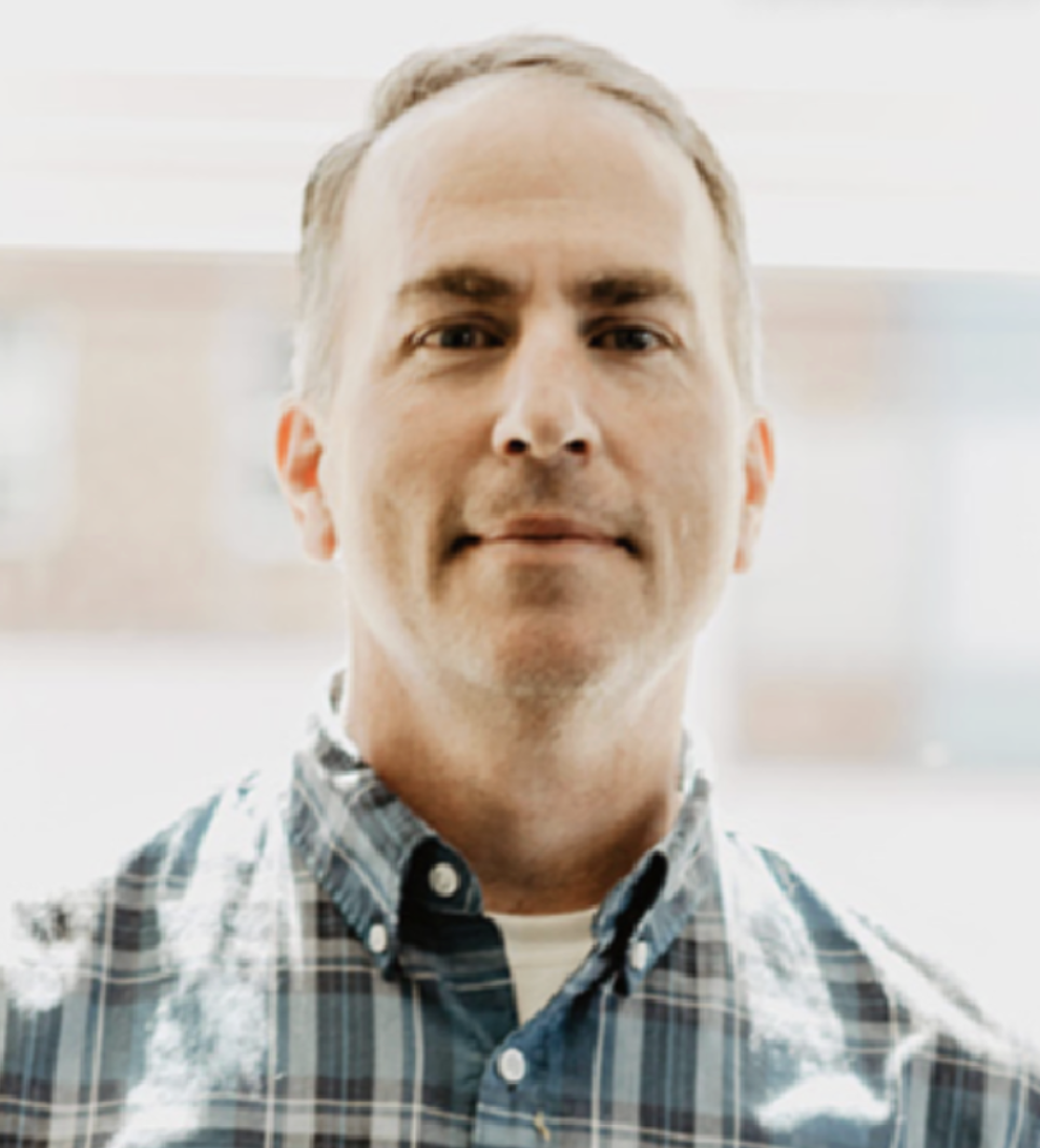 Portrait of a man with short gray hair, wearing a blue and gray plaid button-up shirt, standing indoors with a blurred background.