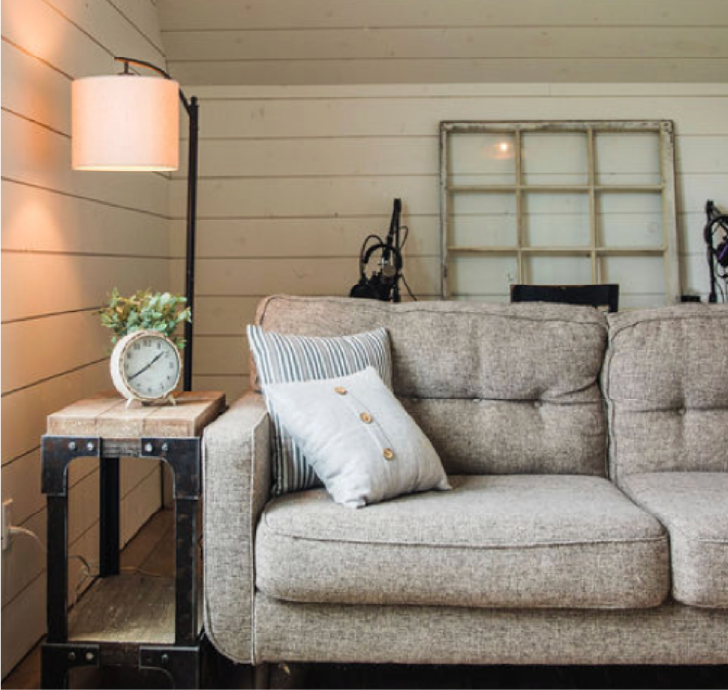Living room with beige fabric sofa, striped and button-tufted pillows, wooden side table with decorative clock and plant, lamp with beige shade, white shiplap wall, and window frame on wall.