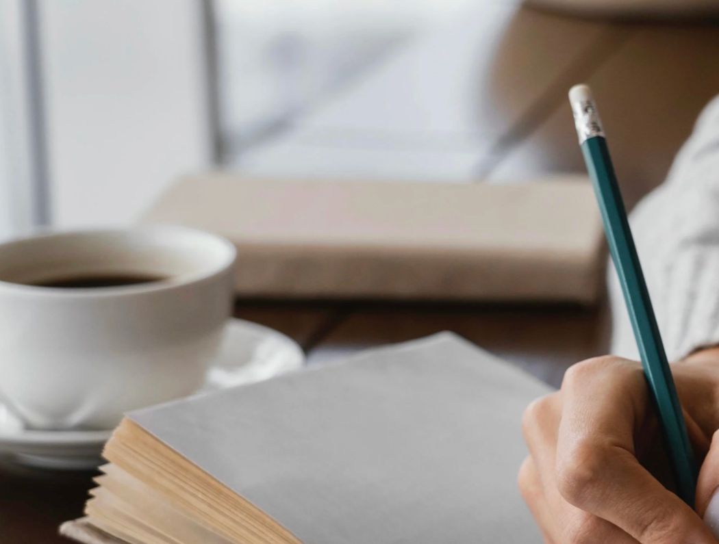Person writing in a notebook with a pencil near a cup of coffee on a desk.