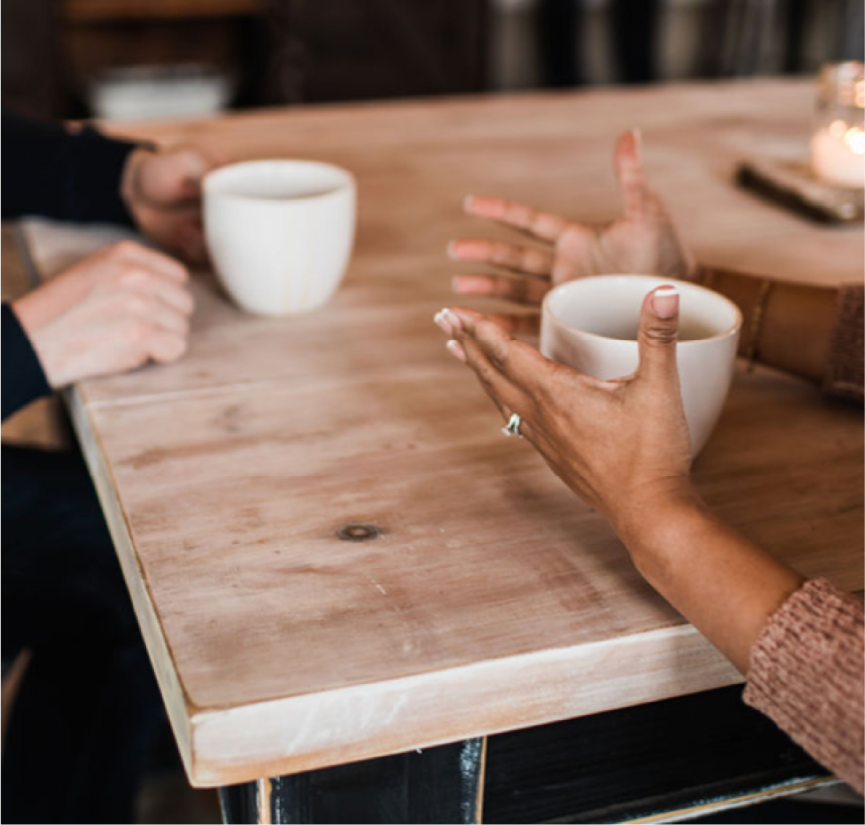 Two people sitting at a wooden table, each holding a coffee mug, engaged in conversation.